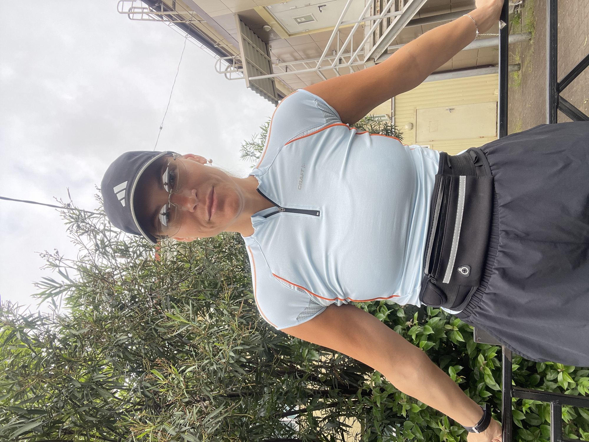 A woman wearing a light blue sports shirt with orange trim, black shorts with a black waist pack, a black baseball cap with a white Adidas logo, and sunglasses is standing outside. She has her hands on a black metal railing. There are green trees and bushes behind her and a light yellow building with metal stairs in the background. The photo was taken in Sochi, Russia.