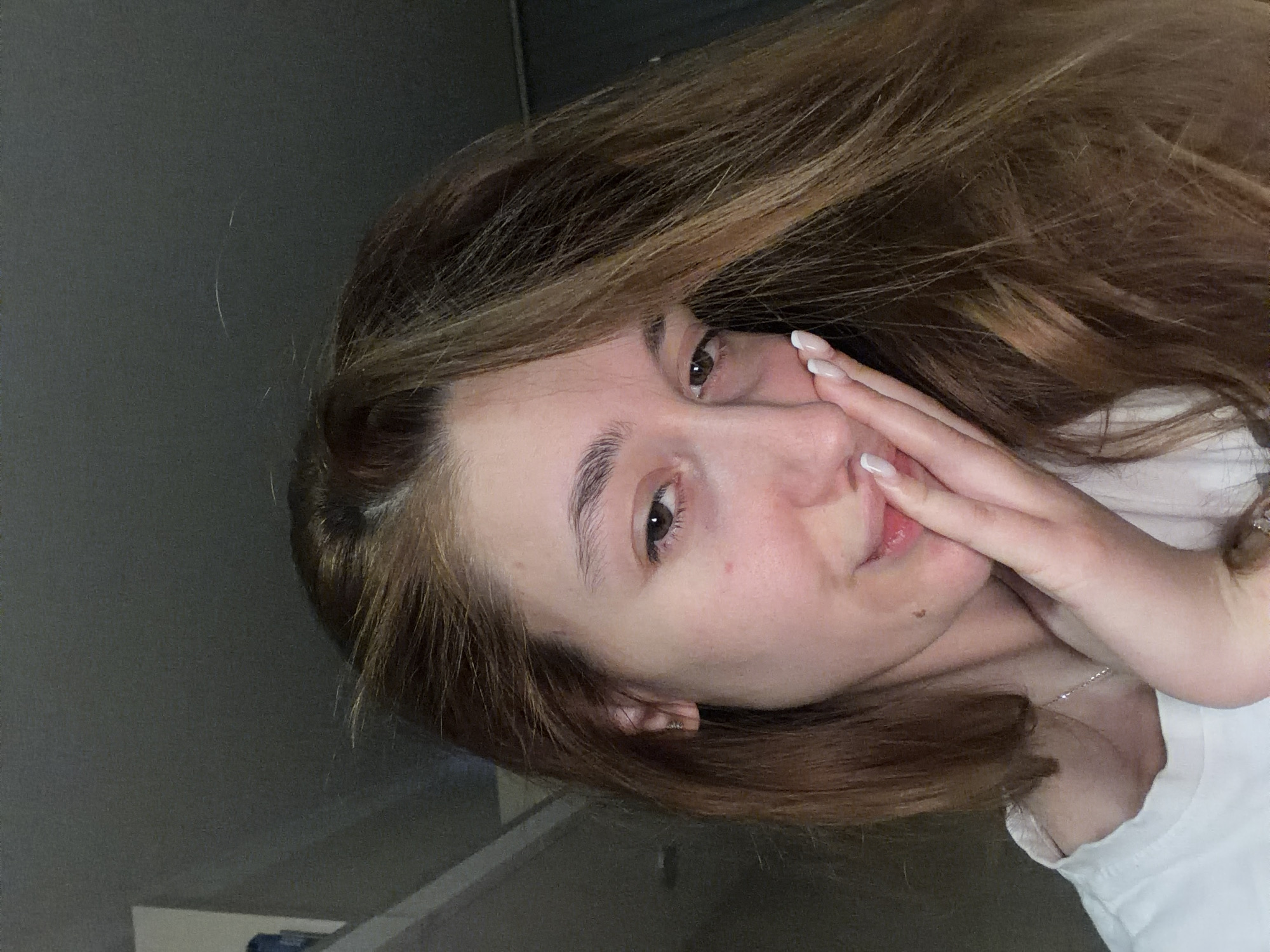 A young woman with light brown hair and fair skin is posing for a selfie. She is wearing a white t-shirt and a delicate silver necklace. Her hair is partially covering her face and one hand is raised to her cheek. She has a slight smile. The background is a plain, gray wall. There is a white object in the bottom left corner, possibly a shelf or piece of furniture. No information is available to determine if this image was taken in Sofia, Bulgaria.