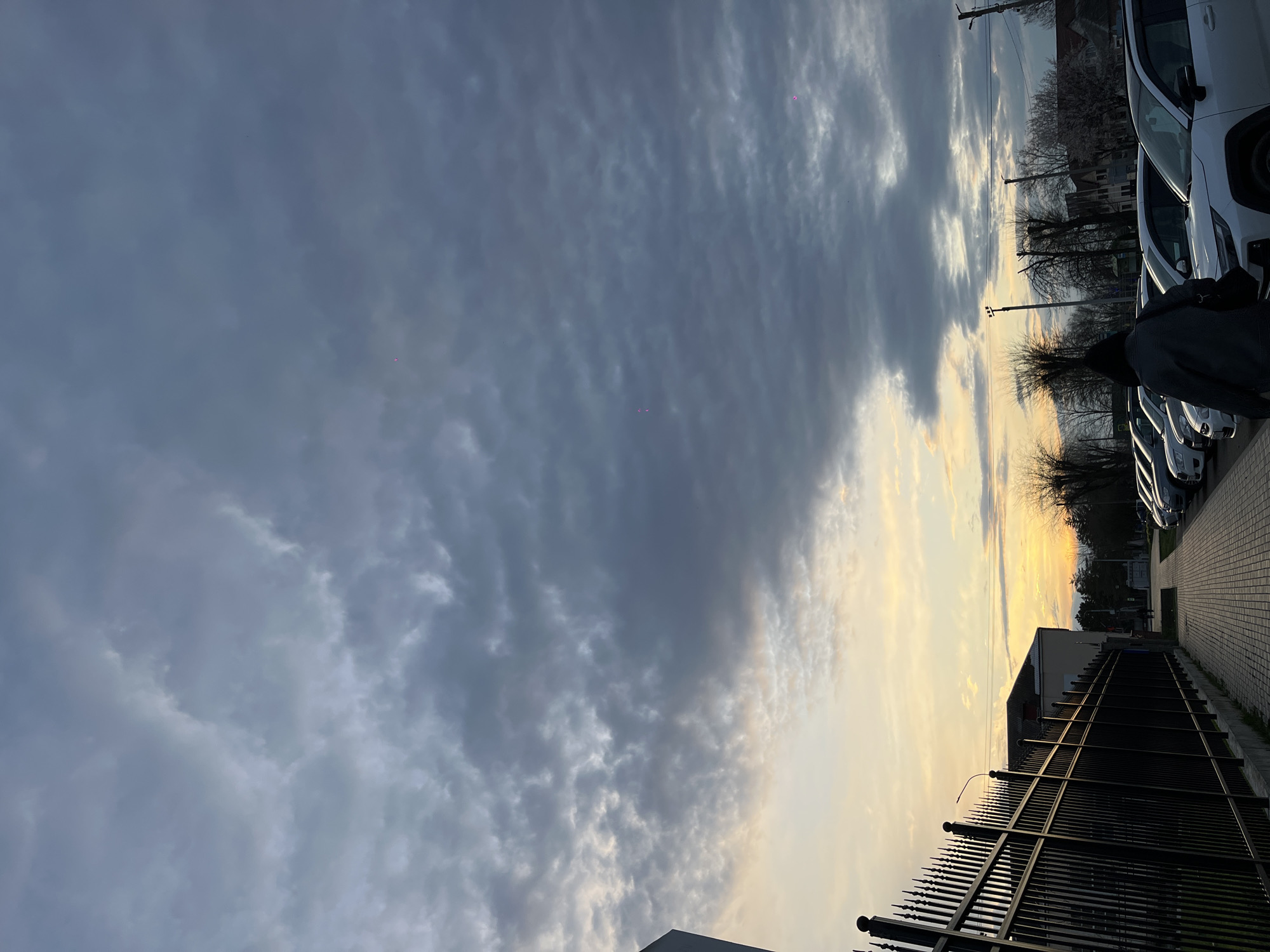 The image shows a view of a cloudy sky with a sunset visible on the right side. Below the sky, there is a street with parked cars on the right and a fence on the left. A person is partially visible near the parked cars. The location appears to be a residential or commercial area in Novopokrovskaya, Russia.
