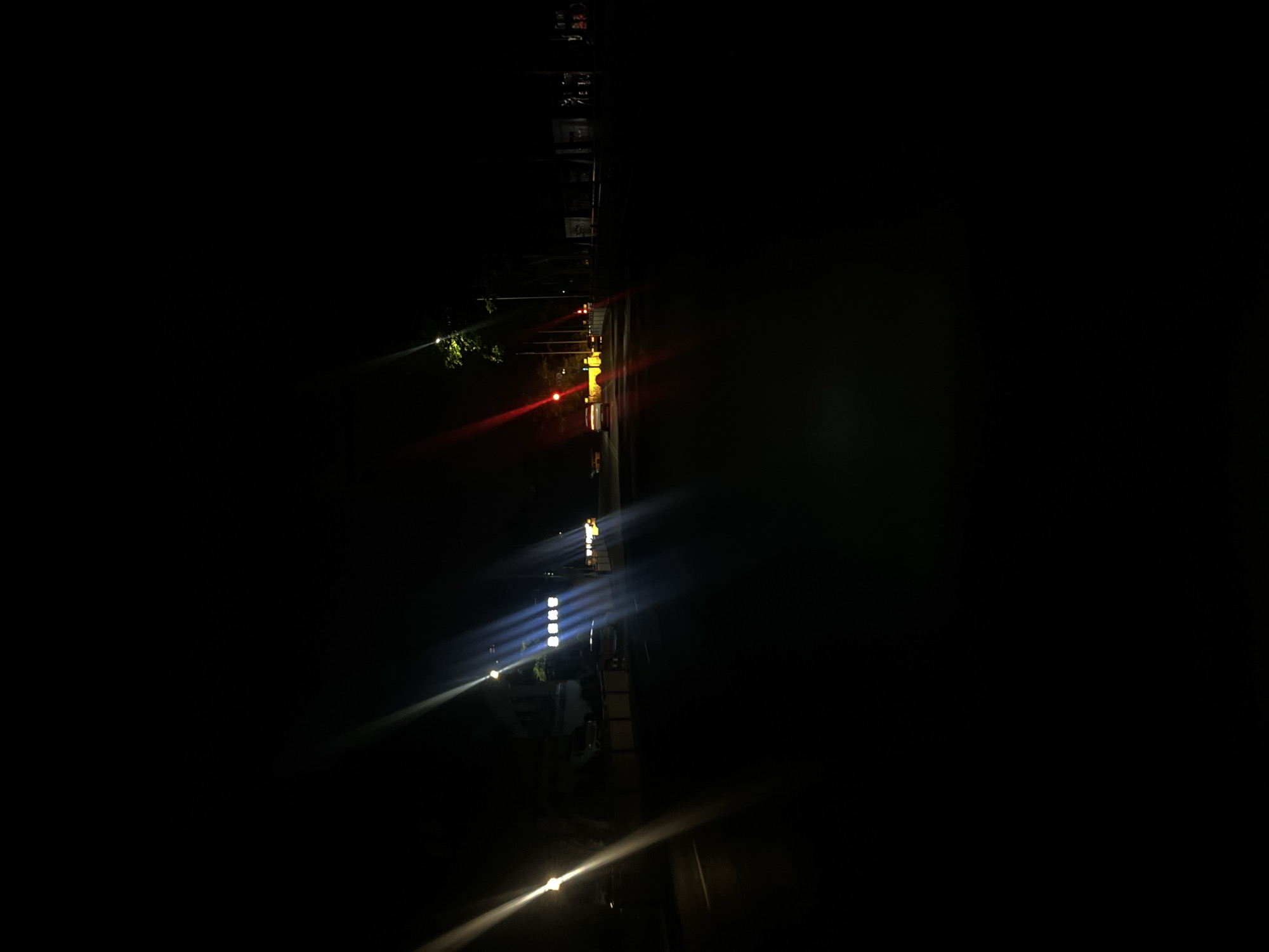 This is a nighttime vertical photograph of an urban street in Jiulongpo District, China. The scene is predominantly dark, illuminated by multiple artificial light sources that produce significant lens flares.
On the far left, a bright, warm-toned streetlight is visible, casting a strong diagonal flare across the lower left portion of the image. A faint white road marking is discernible on the dark asphalt road surface. In the mid-ground on the left, another streetlight of similar luminosity is present.
Towards the center-left, a building features several illuminated square or rectangular fixtures emitting a bright, cool white or blue light. Adjacent to this building, a bright light source, likely a street lamp, generates prominent vertical blue-white lens flares. A dark-colored vehicle, possibly a van or small truck, is parked or stationary near this building.
On the center-right, an active red traffic light is visible, projecting a distinct red diagonal flare downwards and to the left. A light-colored sedan is positioned in the road lane, stopped before the red traffic light. Further right, dark foliage from trees and indistinct urban structures are present, partially illuminated by ambient light.
The sky is completely dark. The road surface is dark asphalt. No specific individuals or explicit actions are clearly visible beyond the implied presence of vehicles on the street.