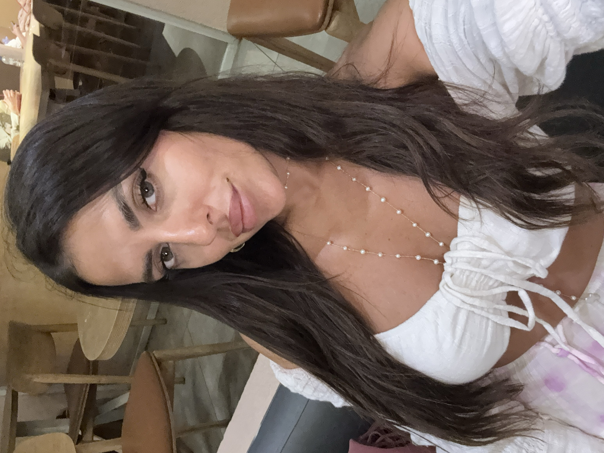 A woman with long, dark, wavy hair is pictured in a close-up, selfie-style shot. She is wearing an off-the-shoulder white top with ruffled sleeves and a string tie in the front. A delicate pearl necklace is draped around her neck. She is also wearing light-colored bottoms with a tie. Her makeup is visible, including eyeliner and mascara. In the background, wooden chairs and tables suggest an indoor seating area, possibly a cafe or restaurant. The environment appears to be dimly lit.