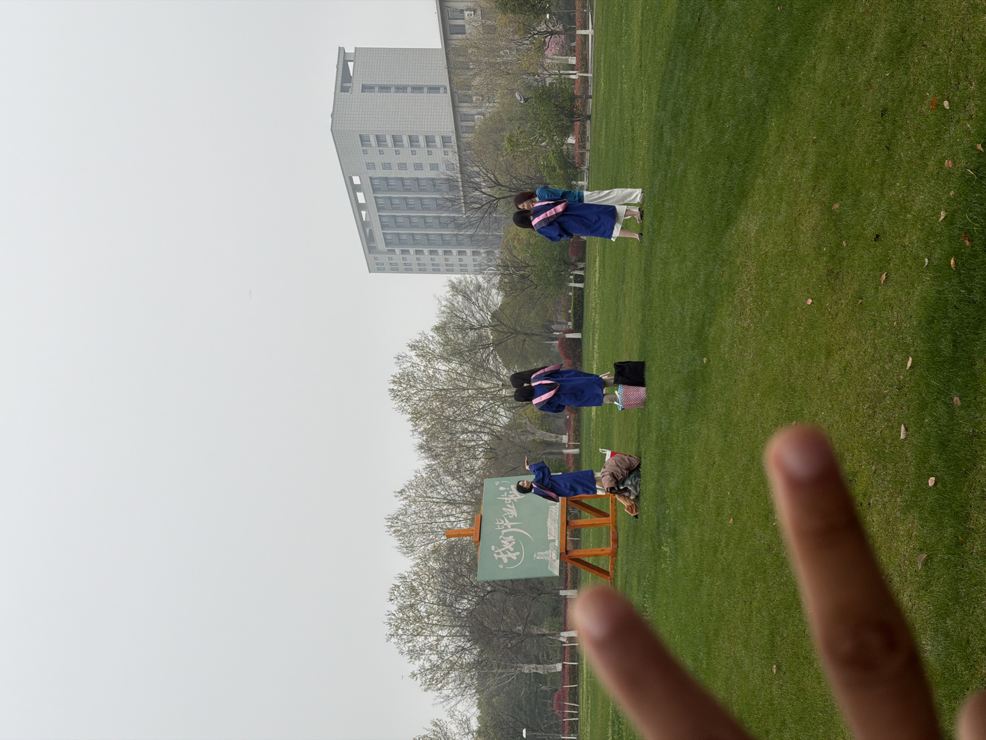The image shows a scene in Jiangning District, China, with a grassy area, a tall building in the background, and trees. Several people wearing graduation gowns are visible; some are interacting with a chalkboard-like structure with writing on it. A pair of fingers is in the foreground, forming a "peace" sign.