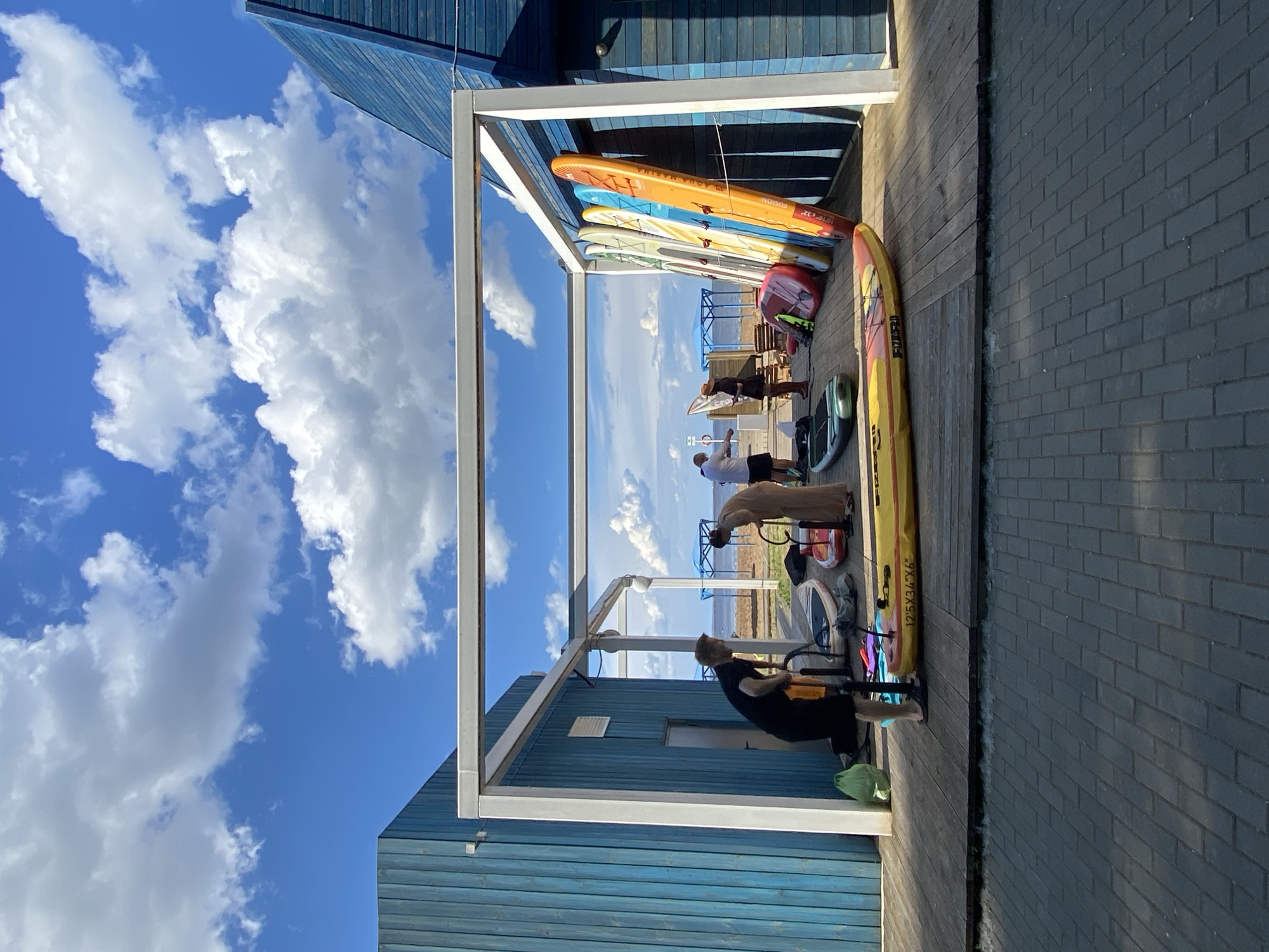 The image depicts a scene on the shore of the Kama River in Chaikovsky, Russia, likely at a water sports rental facility. In the foreground, a wooden deck separates a paved area from the beach. A person in black shorts and a black t-shirt is operating a bright orange hand pump connected to a yellow paddleboard positioned horizontally on the deck. To the right of this person, a second paddleboard lies on its side, red and white. Further back, several other paddleboards of various colors (including orange, yellow, blue, and white) are propped upright against a blue wooden building. Three individuals are visible in the middle ground; one in a tan outfit appears to be bending over a red object on the deck, while two others stand nearby. The building has a modern, angular design with blue horizontal siding and a white frame structure extending outwards. The background reveals the Kama River, a sandy beach, and distant blue umbrellas. The sky is a vibrant blue with scattered white cumulus clouds.