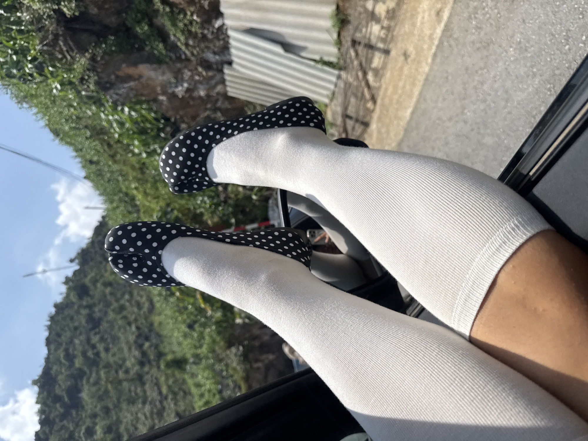 The image shows a pair of legs wearing white knee-high socks and black and white polka-dotted flats extending from a car window. The background includes greenery, a hillside, and a corrugated metal structure. The location is Lũng Phìn Commune, Vietnam.