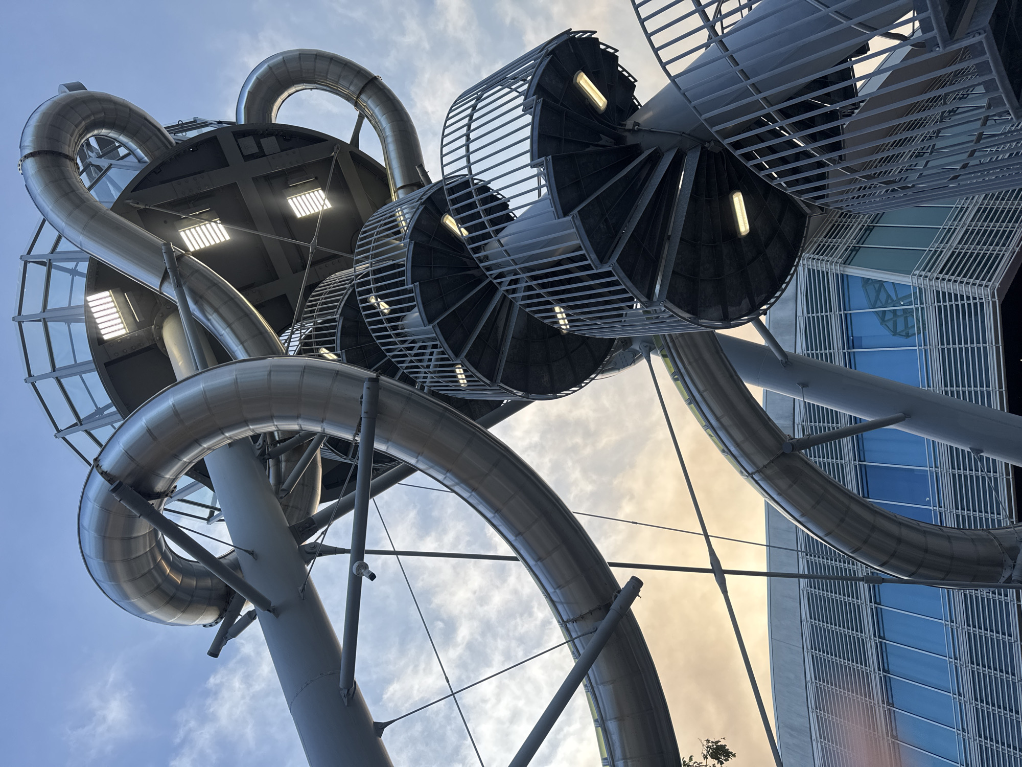 An upward, low-angle shot captures a sliver of a steel slide structure against a cloudy sky. The structure is composed of large, curved, silver tubes, reminiscent of a DNA helix, connected by spiral staircases and platforms. The tubes twist and turn, creating a dynamic, almost abstract form. One of the spiral staircases, made of dark metal with a railing, is prominently featured in the foreground. The staircase has several landings and is illuminated by rectangular lights. In the background, a modern building with a facade of horizontal metallic louvers and blue-tinted windows is visible. The sky above is a mix of blue with scattered white clouds, and a hint of golden sunset light filters through the clouds near the horizon. The overall impression is one of architectural ingenuity and playful design within an urban setting.