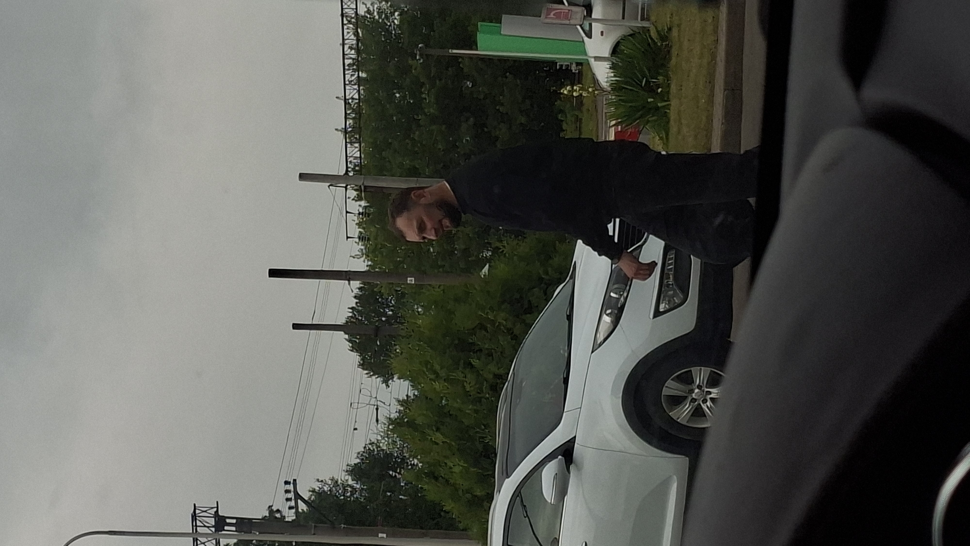 A man in a black long-sleeved shirt and dark pants walks near a white SUV on a cloudy day in Smalyavichy, Belarus. The man is facing left and has a beard. He is walking on a paved surface next to the SUV, which is parked. In the background, there are trees, utility poles, and overhead wires. A portion of another vehicle is visible in the foreground.