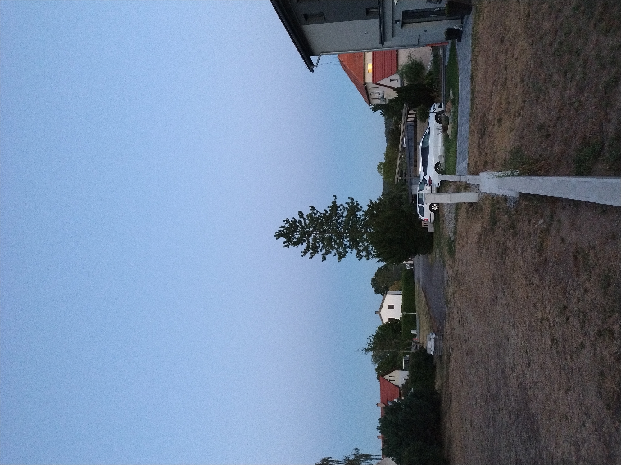 This is an outdoor, vertical, full-shot photograph taken from ground level of a residential area. The sky is a clear, pale blue, suggesting it is either early morning or late evening. In the foreground, a dry, brown grassy lawn is visible, bisected by a low, light-colored concrete wall. Beyond the wall, a paved pathway leads to a driveway where two white cars are parked: a smaller hatchback and a larger wagon. To the left of the cars, a white minivan is parked in front of a building with a red-tiled roof. Further left, several evergreen trees and shrubs are seen.
In the middle ground, a tall, slender evergreen tree stands prominently. Behind it, a dark asphalt road curves away, bordered by more greenery. Several houses are visible, including a white house with a red-tiled roof and a white house with a dark roof. To the far right, a multi-story building with a gray facade and a dark-tiled roof dominates the edge of the frame, featuring a visible gutter and downspout.
The image appears to be located in the city of Dobev, Czechia, based on the architectural style of the houses and the overall environment. There are no people visible in the scene.