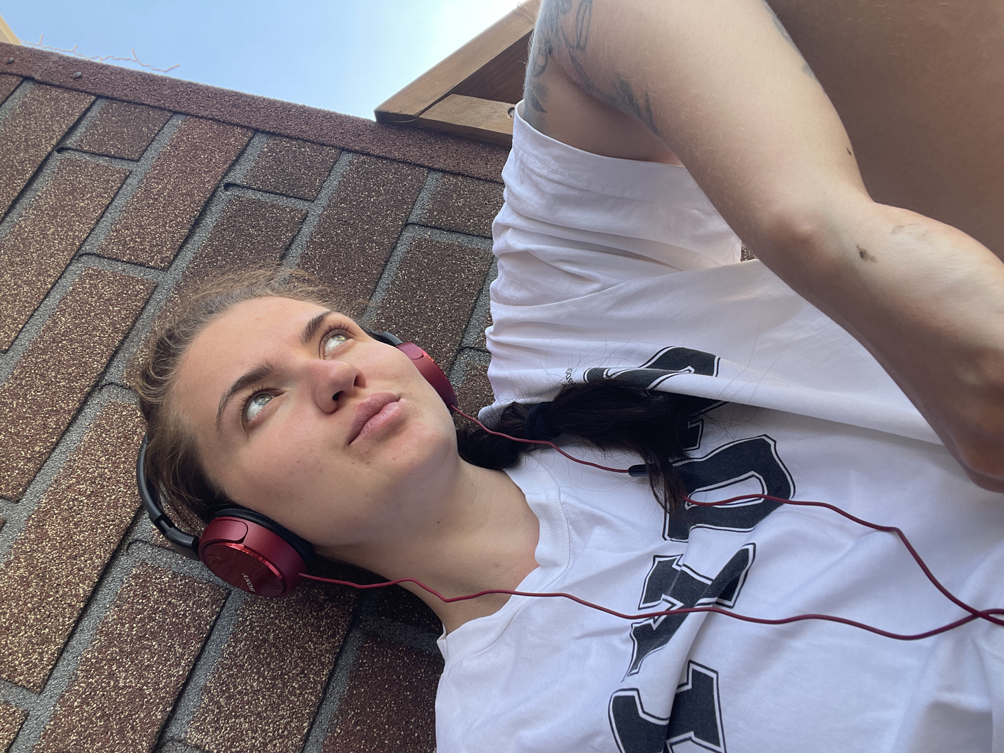 A young woman with fair skin is lying on a brown asphalt shingle roof, wearing red Sony headphones and a white t-shirt with black letters. She is looking up and slightly to the left. Her dark hair is pulled back. Part of her tattooed arm is visible. The sky is light blue. The location is Мосметаллоконструкция Онт, Russia.