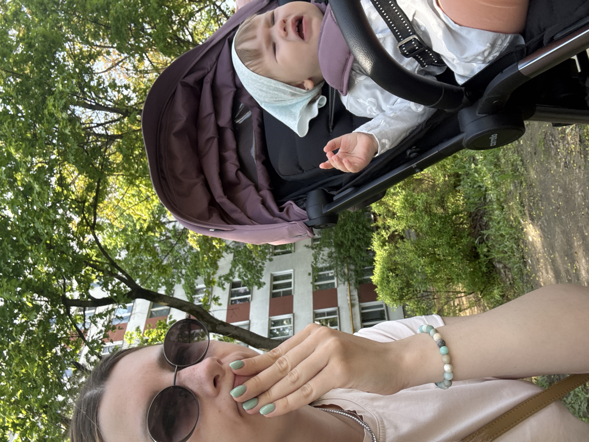 A downward-angled, personal photograph shows a woman in sunglasses holding a hand to her mouth in a possible gesture of exasperation or silencing, with a baby crying in a stroller above her. The baby is light-skinned, wearing a white garment and light blue head covering, situated in a plum-colored stroller. The woman is light-skinned with a beaded bracelet visible on her wrist. Behind them is a light-colored building with multiple windows, characteristic of urban architecture. Green trees are also present. The photo appears to have been taken outside in a residential area. Given the urban architecture style, the photo might have been taken in Moscow, Russia.