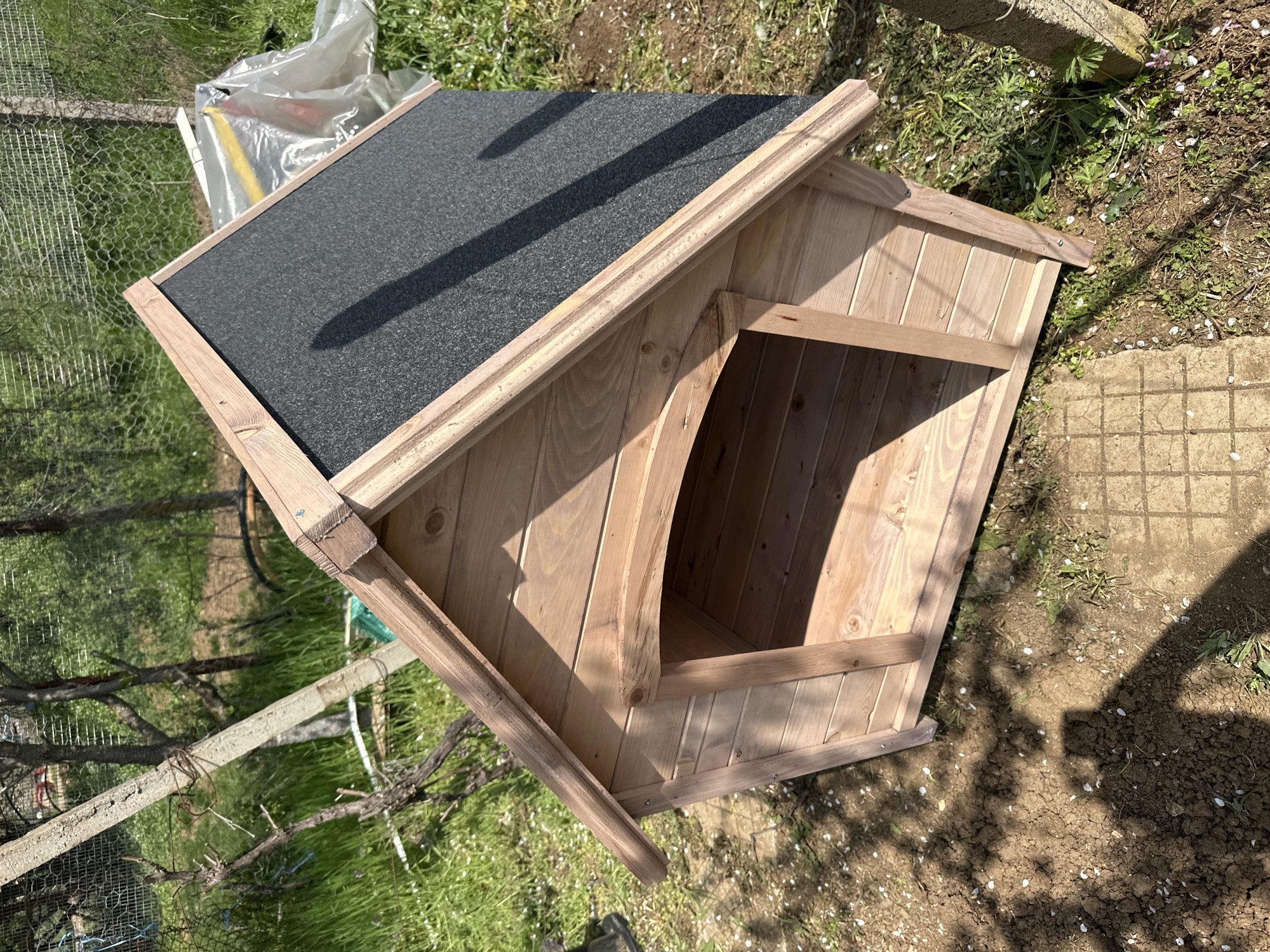The image shows a newly constructed wooden doghouse with a black shingled roof sitting on the ground in a grassy area. The doghouse has an arched opening. The location appears to be outdoors, with signs of greenery and soil. There is a wire fence and a concrete post visible in the background. The image does not contain people. There is no indication of the city Montana, Bulgaria.