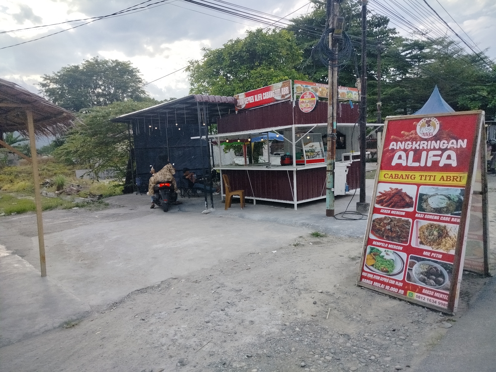 The image depicts an outdoor food stall named "Angkringan Alifa Cabang Titi Abri" in Kampungmelayu Satu, Indonesia. A red A-frame sign in the foreground displays various food items offered, including "Ceker Mercon," "Nasi Goreng Cabe Raw," "Rempelo Mercon," "Mie Petir," and "Bakso Mentel." The food stall itself is a modest structure with a dark brown roof and white framing, topped with signage that reads "AYAM GEPREK ALFA CABANG TITI ABRI." To the left of the stall, a thatched roof structure, possibly a waiting area or additional seating, is visible. A motorcycle is parked near the stall with a person seated on it. Several tables and chairs are arranged under the dark roofed section of the stall. The ground is a mix of concrete and rough gravel. In the background, a dense canopy of trees forms a green backdrop under an overcast sky. Electrical wires are strung across the upper portion of the image.