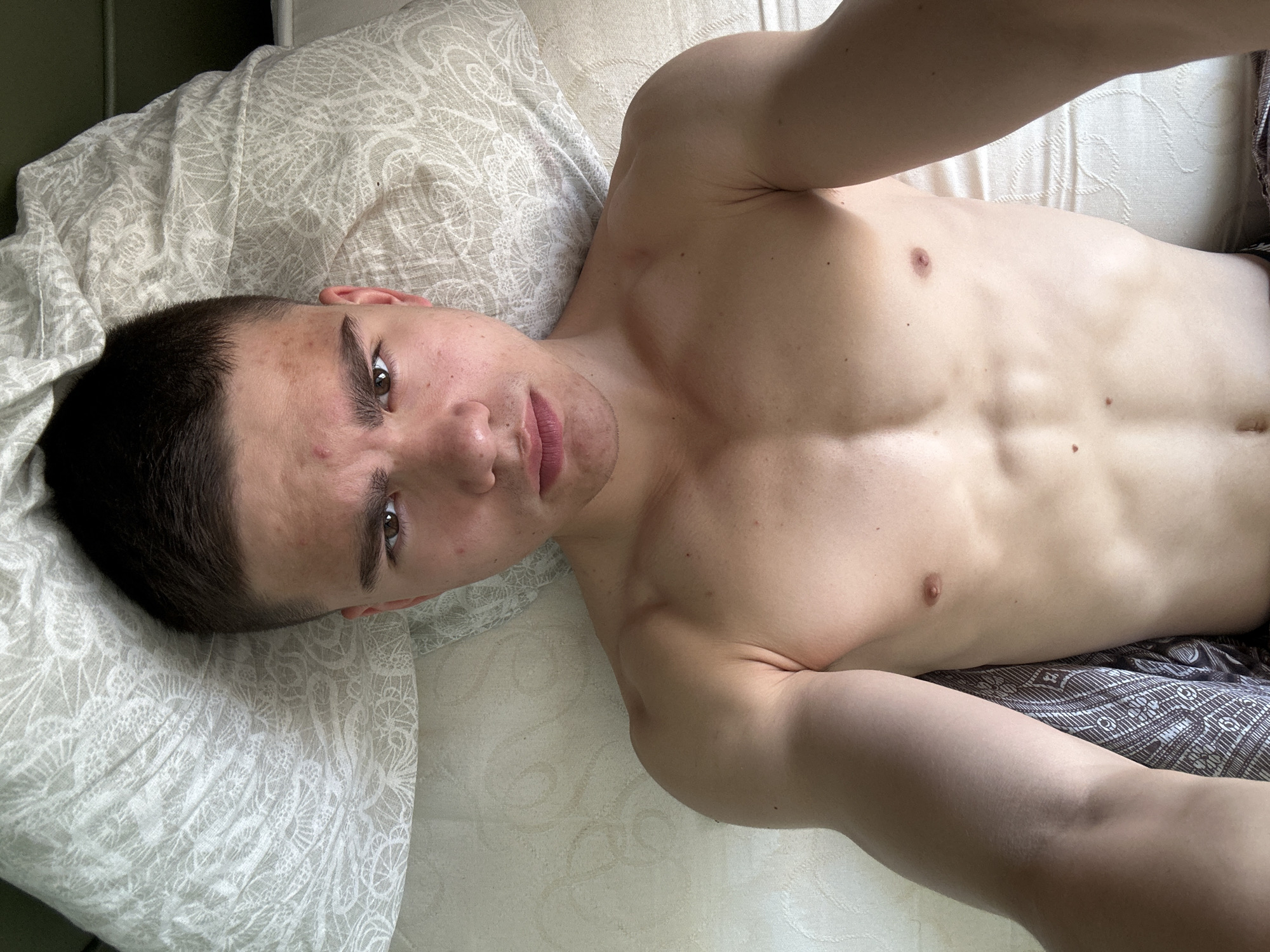 A medium shot shows a shirtless male with short dark hair, lying on his back on a bed with a patterned pillow and comforter. The location is an interior, city Unknown, Unknown. The male faces the camera with a neutral expression. Visible physical features include muscular chest and shoulders, and some acne on the face. His arms are extended, presumably holding a camera for a selfie. The background is slightly out of focus.