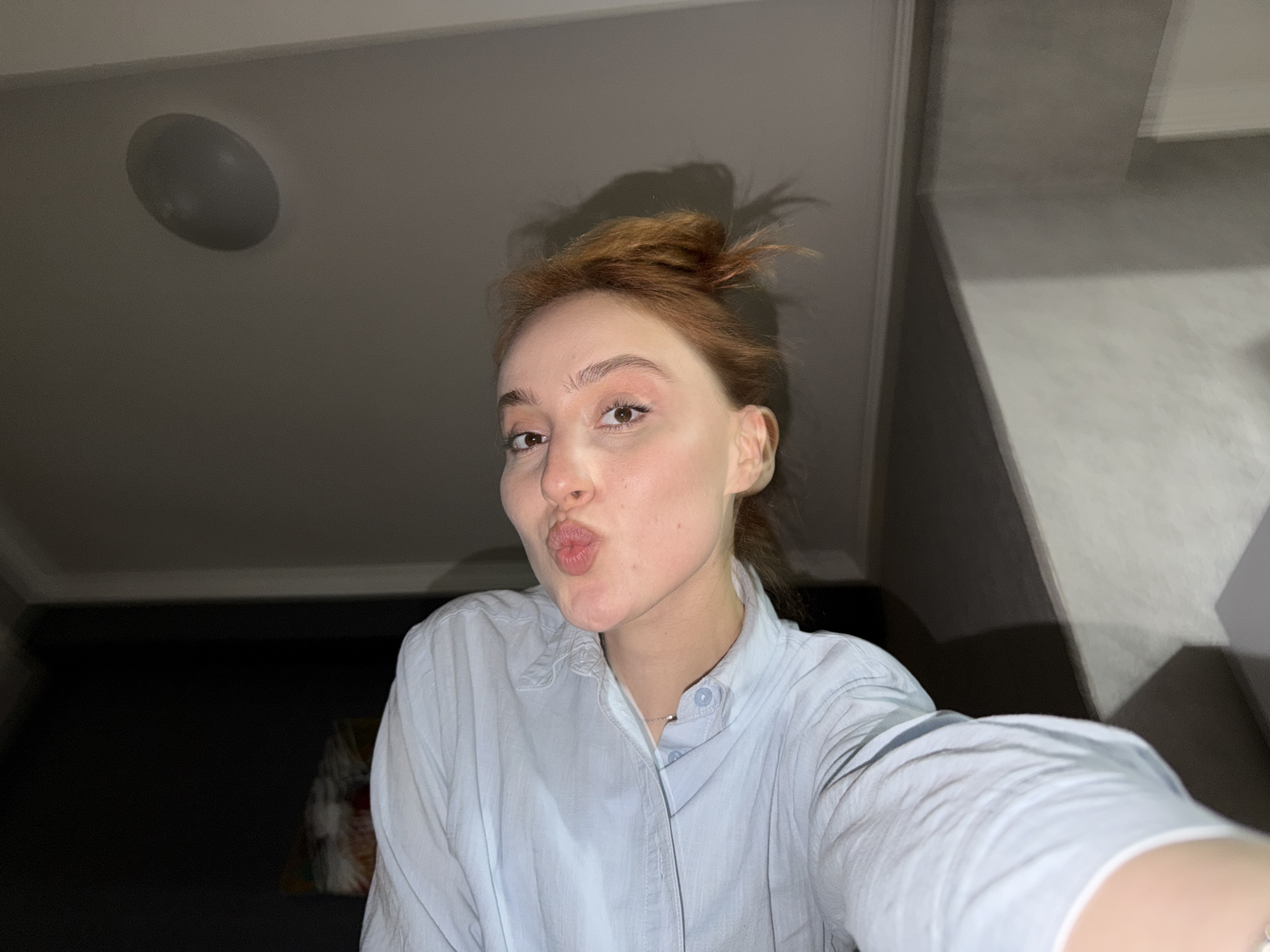 A selfie of a fair-skinned woman with reddish-blonde hair pulled into a messy bun. She is wearing a light blue button-down shirt and making a kissing face. The background appears to be an interior room with a ceiling fixture and indistinct dark objects. There's no evidence to pinpoint this picture to Tbilisi, Georgia.