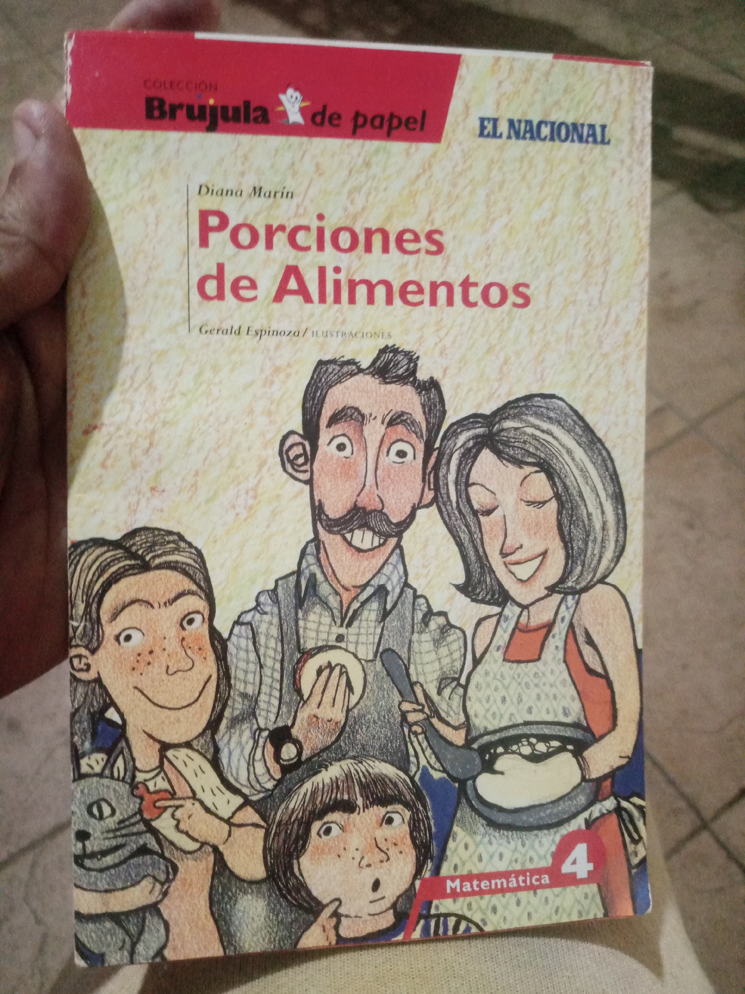 A hand holds a book titled "Porciones de Alimentos" (Food Portions) by Diana Marin, with illustrations by Gerald Espinoza. The book features a colorful illustration of a family preparing food, including a father with a mustache, a mother in an apron, and two children. A cat is also visible in the illustration. The book is part of the "Brujula de papel" collection and is published by "EL NACIONAL." The book has a red corner with the number "4" and the word "Matemática" (Mathematics). The background is a textured, light-colored surface. The image appears to be taken indoors, possibly in Santa Ana, Venezuela, given the book's publisher.