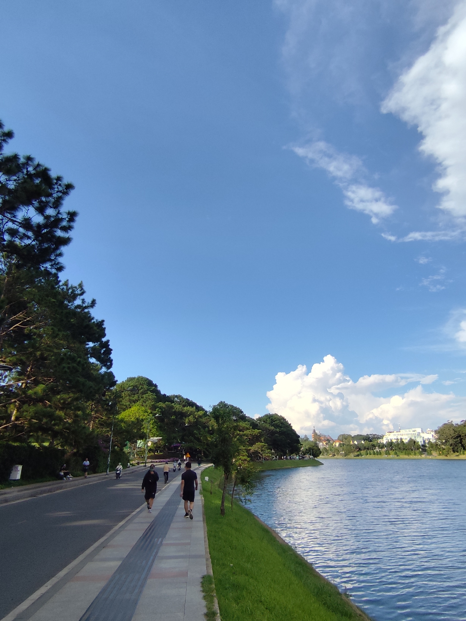 This image depicts a scenic view of Xuan Huong Lake in Da Lat, Vietnam. The foreground features a paved walkway alongside the lake, bordered by lush green grass. Two individuals are walking on the path, heading away from the camera. A road runs parallel to the walkway, with several people and a few motorcycles visible. The lake itself occupies the right side of the frame, with gentle ripples on its surface reflecting the bright blue sky. Along the far bank of the lake, a line of trees creates a natural barrier, beyond which are buildings with white facades and some with visible architectural details, suggesting a developed urban area. The sky is a clear, bright blue with scattered white clouds. The overall scene is tranquil and suggests a pleasant day for outdoor activity.