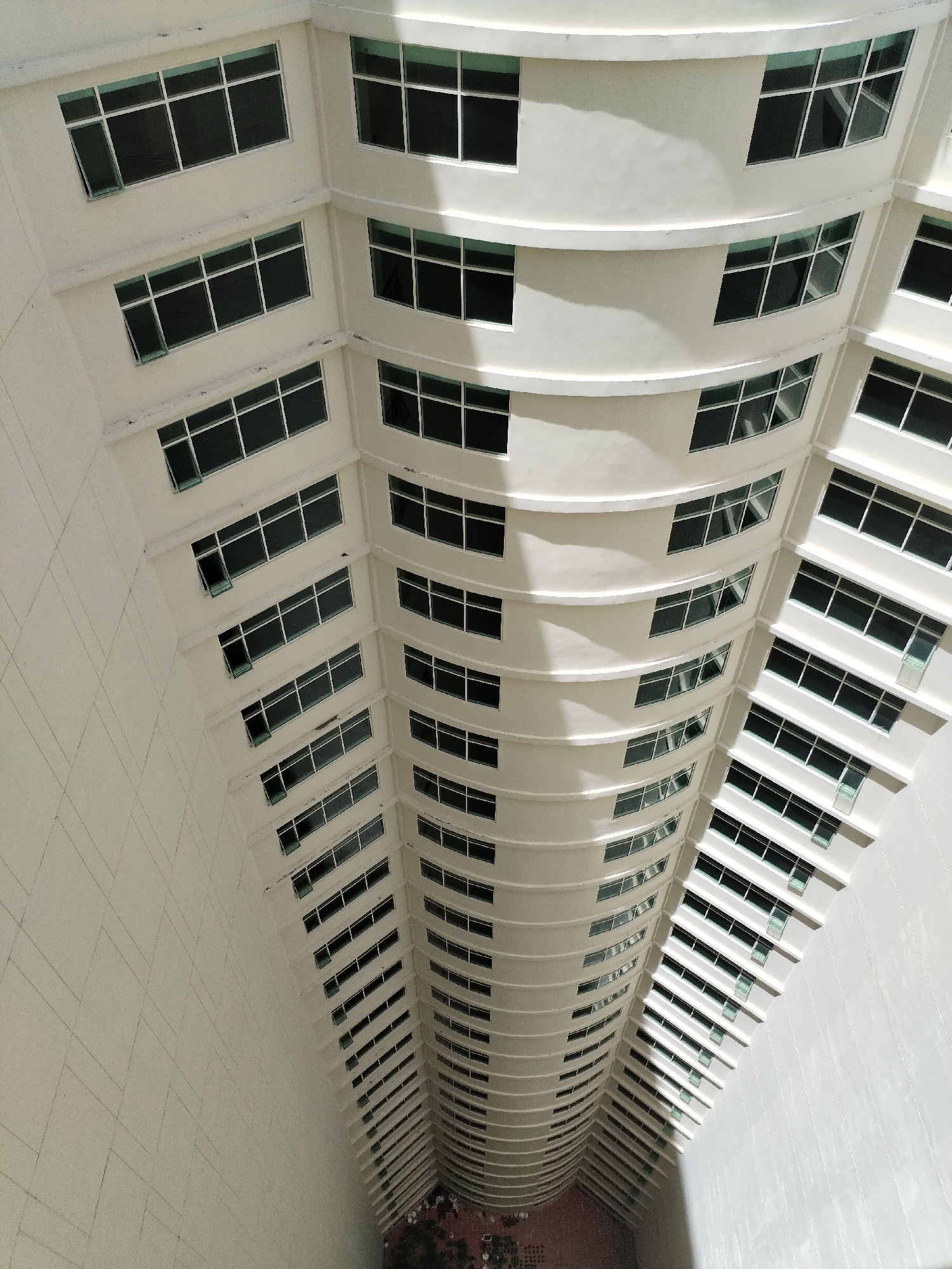 The image is a high-angle, downward shot of the exterior of a modern, white, multi-story building, likely a hotel or residential complex. The building has a curved facade with numerous windows, each divided into multiple panes. The windows are dark, reflecting the sky or interior. Sunlight casts shadows on the building, highlighting its architectural details, such as the protruding window frames and horizontal ledges. The scene appears to be in an urban environment, given the scale of the building, but the specific location of Nha Trang Ward, Vietnam is not visually discernible from the image itself. There are no people visible in the frame. The overall impression is one of clean, contemporary architecture against a bright sky.