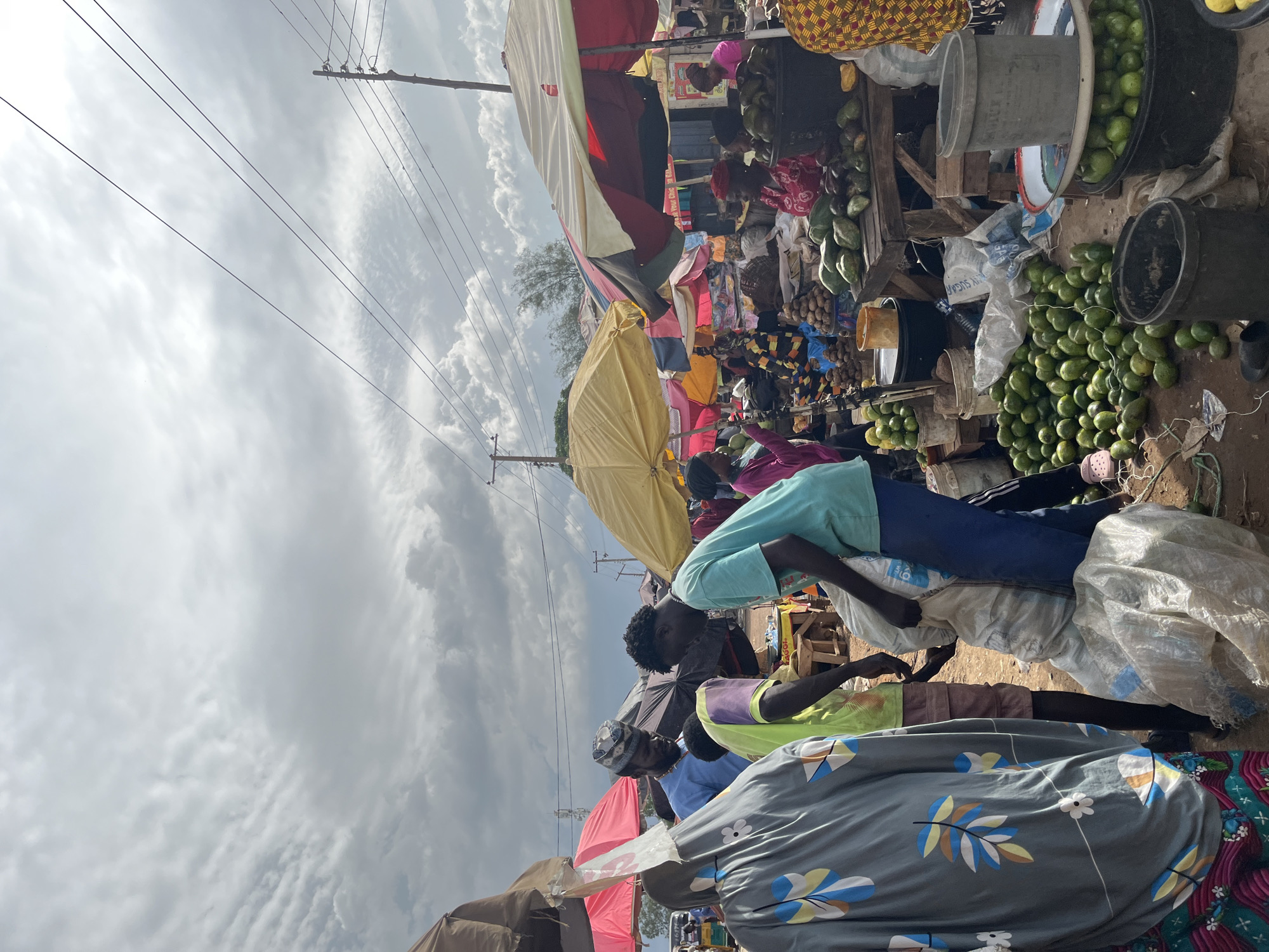 A busy outdoor market scene is captured under a cloudy sky in Rafin Par, Nigeria. The foreground is dominated by a large pile of green avocados on the dirt ground, with several other containers and baskets filled with avocados also visible. To the left, a person in a grey, patterned garment stands beside a large white sack. In the center, two men are interacting near the sack. One man, wearing a light blue shirt and a patterned cap, is looking down, while another man in a turquoise t-shirt is holding the sack. Behind them, various stalls are set up with colorful umbrellas, displaying more produce and goods. Several people are milling about the market, engaged in shopping and selling activities. Power lines are visible against the sky, indicating the presence of infrastructure within the urban setting.