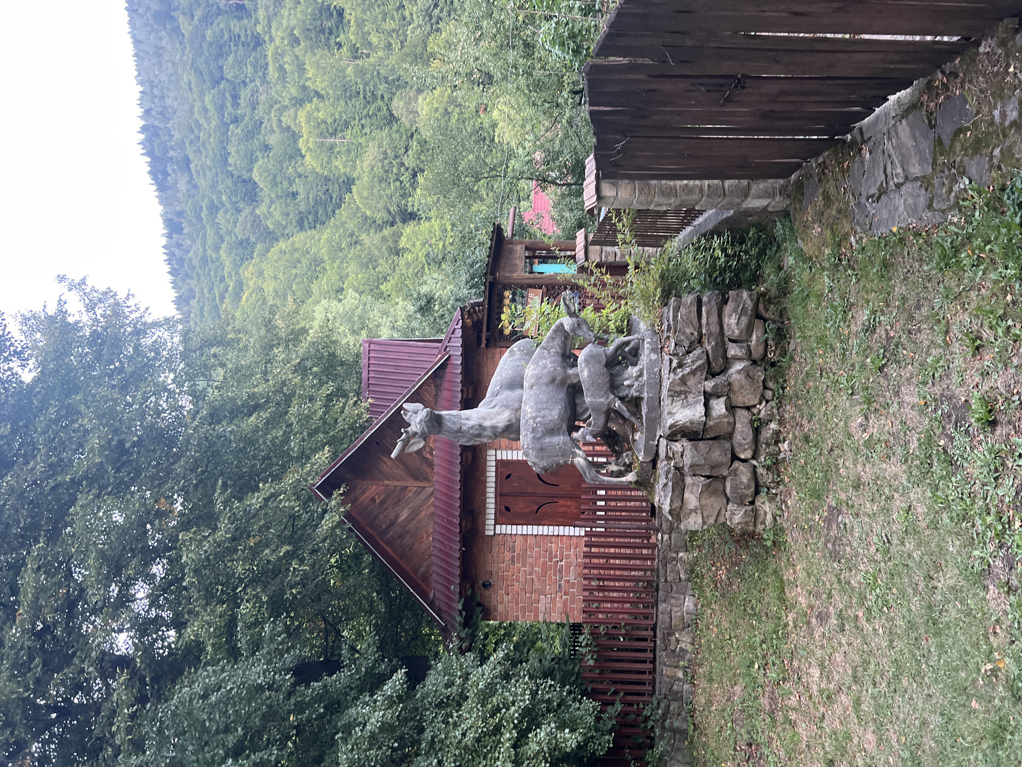 The image depicts a scenic outdoor view in Yaremche, Ukraine, focusing on a stone sculpture of deer in front of a rustic brick building. The sculpture features a larger deer with antlers, a smaller deer standing next to it, and a fawn positioned at the base. The building behind the sculpture is constructed of red brick with a wooden roof and wooden double doors. A wooden fence borders the property. The environment is lush and green, with dense trees and foliage surrounding the building and extending up a hillside in the background, suggesting a natural, possibly mountainous or forested setting. The lighting suggests it is daytime, with overcast skies.