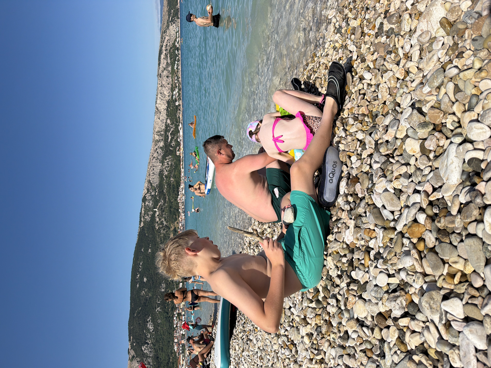This media file depicts a sunny day at a pebble beach in Općina Baška, Croatia. Several people are enjoying the water and the shore.
In the foreground, a young boy with blonde hair sits on the pebbles, looking to his right and holding a stick. He is wearing green swim shorts. To his right, a man is seated, also on the pebbles, with his arm around a young girl who is wearing a pink bikini with a leopard print pattern and a patterned swim cap. The man is wearing dark swim shorts. Their feet are visible, with the man wearing black water shoes and the girl wearing black swim shoes. A pair of grey water shoes with "AQUOS" visible on the side are also in the foreground.
In the background, the turquoise sea stretches out, with a mountainous, green landscape and buildings lining the coast. Several people are in the water, some on inflatable rafts. One man is standing in the water, holding a ball. The bright sunlight suggests it is daytime, and the clear blue sky indicates fair weather. The overall mood appears relaxed and summery, with people engaging in typical beach activities.