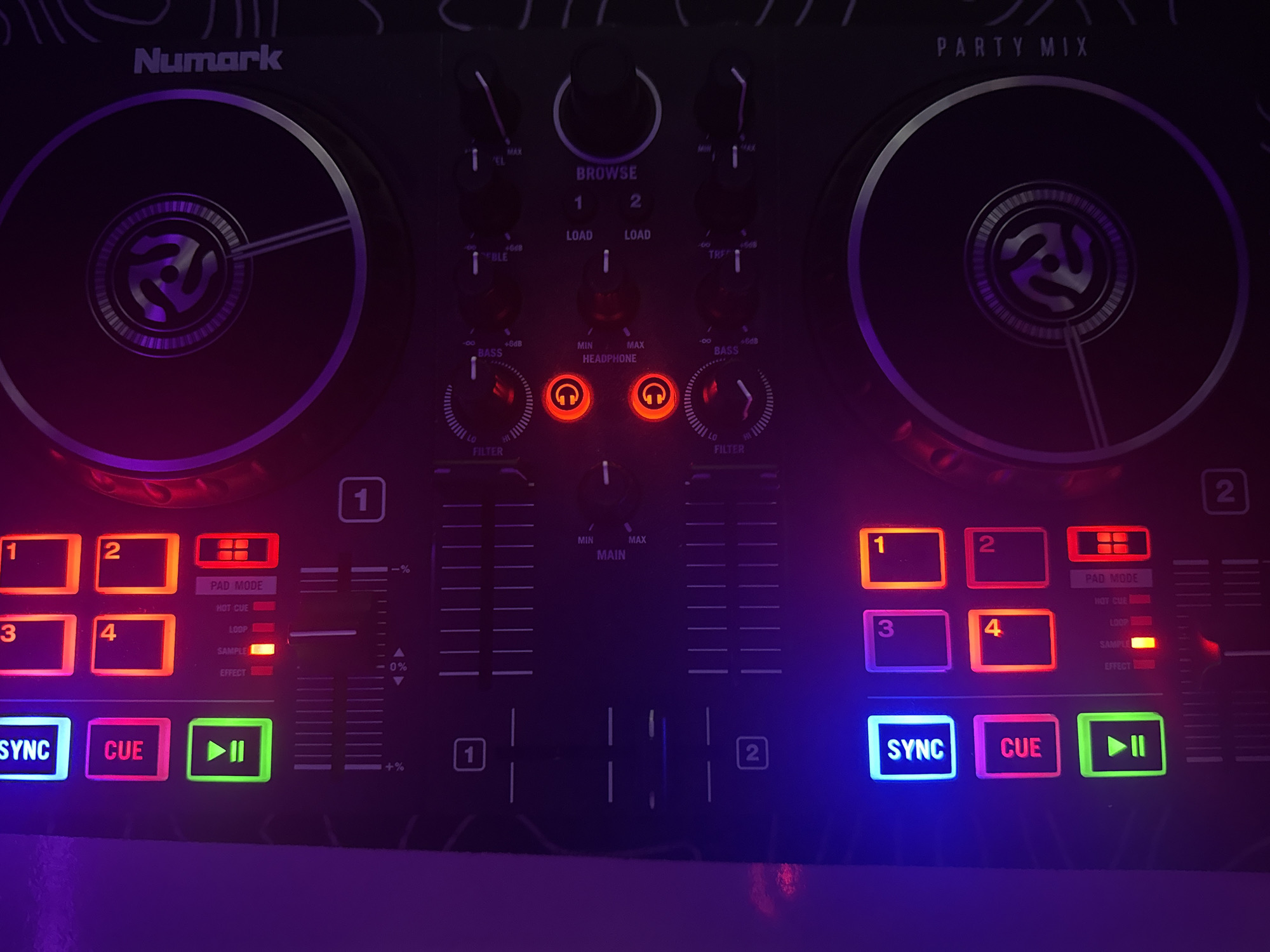 The image shows a close-up view of a Numark Party Mix DJ controller. The controller is dark, with various buttons and knobs illuminated by colorful LED lights, primarily blue, pink, red, and orange. The jog wheels are prominently featured, with the Numark logo visible on them.
Specifically, the image shows:
* **Top Left:** A blue illuminated "SYNC" button and a pink illuminated "CUE" button. Below these are four square buttons, two illuminated in orange (#3 and #1) and two in red (#4 and #2). A green illuminated play/pause button is below the CUE button, with a red indicator light next to a "LOOP" label.
* **Right Side:** The large jog wheel with the Numark logo. Text on the right side reads "PARTY MIX".
* **Center:** Various knobs and sliders. Some are illuminated red, such as two "HEADPHONE" volume knobs. Other controls are labeled "FILTER," "BASS," "TAP," "LOAD," and "BROWSE." A circular knob labeled "BROWSE" is visible on the right side, with "1" and "2" indicators below it.
* **Bottom Left:** Another set of buttons and controls, including a blue illuminated "SYNC" button and a pink illuminated "CUE" button. Below these are two illuminated square buttons, one in orange (#3) and one in red (#4). A green illuminated play/pause button is visible below the CUE button, and a red indicator light is next to a "LOOP" label. A vertical slider is located below these controls.
The lighting is primarily from the DJ controller itself, with some ambient purple and blue light reflecting on the surface. The background is dark and out of focus, suggesting the image was taken in a dimly lit environment. There are no people visible in the image.
The location in the city of Burgas, Bulgaria, is not determinable from the provided image.