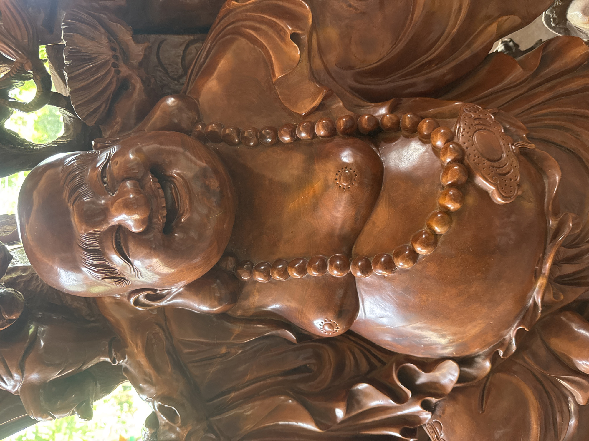 The image presents a detailed, frontal view of a large, meticulously carved wooden statue of Budai, commonly known as the Laughing Buddha. The figure is rendered in a warm, reddish-brown wood, exhibiting a high polish or lacquer finish that reflects light. The statue depicts a robust, smiling figure with a wide, open-mouthed grin, expressive crinkled eyes, and short, stylized hair. A prominent feature is the figure's bare, large belly, across which a necklace of large, round wooden beads is draped. Each nipple is distinctly carved with a circular pattern of dots surrounding a central disc. At the lowest point of the beaded necklace, a flattened, ornate pendant, carved with a cloud-like or auspicious design, rests on the belly. The figure's body is partially obscured by intricately carved folds of drapery, suggesting robes, which blend into the surrounding wooden structure. In the soft-focus background, blurred green foliage and bright ambient light are visible, indicating an outdoor or semi-outdoor environment. Other carved wooden elements, possibly parts of a larger composition or decorative framework, are also visible behind and to the sides of the main figure. The specific location in the city of Bản Đêu, Vietnam, cannot be determined from the visual information provided.