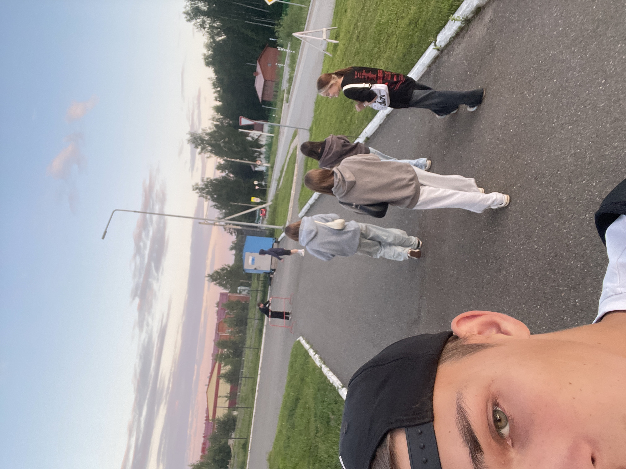 The image depicts a group of young individuals on an asphalt pathway in what is described as Noviy, Russia, during what appears to be either sunset or sunrise, indicated by the pink and purple hues in the sky. In the immediate foreground, the upper left portion of a young male's face is visible, identifying him as the photographer. He wears a black baseball cap reversed, with his left eye, eyebrow, and ear visible. His eye is green. Extending into the mid-ground, five individuals walk away from the photographer on the asphalt path. From left to right: 1. A female in a light grey hooded sweatshirt and matching light grey sweatpants, carrying a white handbag on her left shoulder. 2. A female in a beige hooded sweatshirt and light-colored wide-leg pants. 3. A female in a dark brown or mauve hooded sweatshirt and light pants. 4. A male in a dark blue or black hooded sweatshirt and dark shorts, positioned slightly further back and to the right of the path. 5. A female in a black t-shirt with a red and black graphic design on the back, dark pants, and a small white handbag on her right shoulder. Her blonde hair is pulled back, and she is looking over her left shoulder towards the other individuals or the photographer. The asphalt path features a white painted curb separating it from a green grassy verge on the right. Beyond the verge, a paved road runs parallel, with various road signs, including a white triangular yield sign with a red border and a yellow diamond-shaped sign. A white A-frame barrier stands on the grassy verge to the far right. In the background, a light blue portable cabin is visible on the grassy area to the left of the main road. Near this cabin, two additional individuals are present. One, dressed in dark clothing, is leaning on a red metal barrier. Another individual, also in dark clothing, is partially obscured by the blue cabin. Further in the background, a fence separates the grassy area from several industrial-style buildings with red roofs and yellow or beige walls. One building features a large open bay. Further right, a small, dark red building with a red roof is visible. Numerous trees form a dense treeline behind the structures and along the road. A tall metal streetlight pole is visible on the left side of the path, with