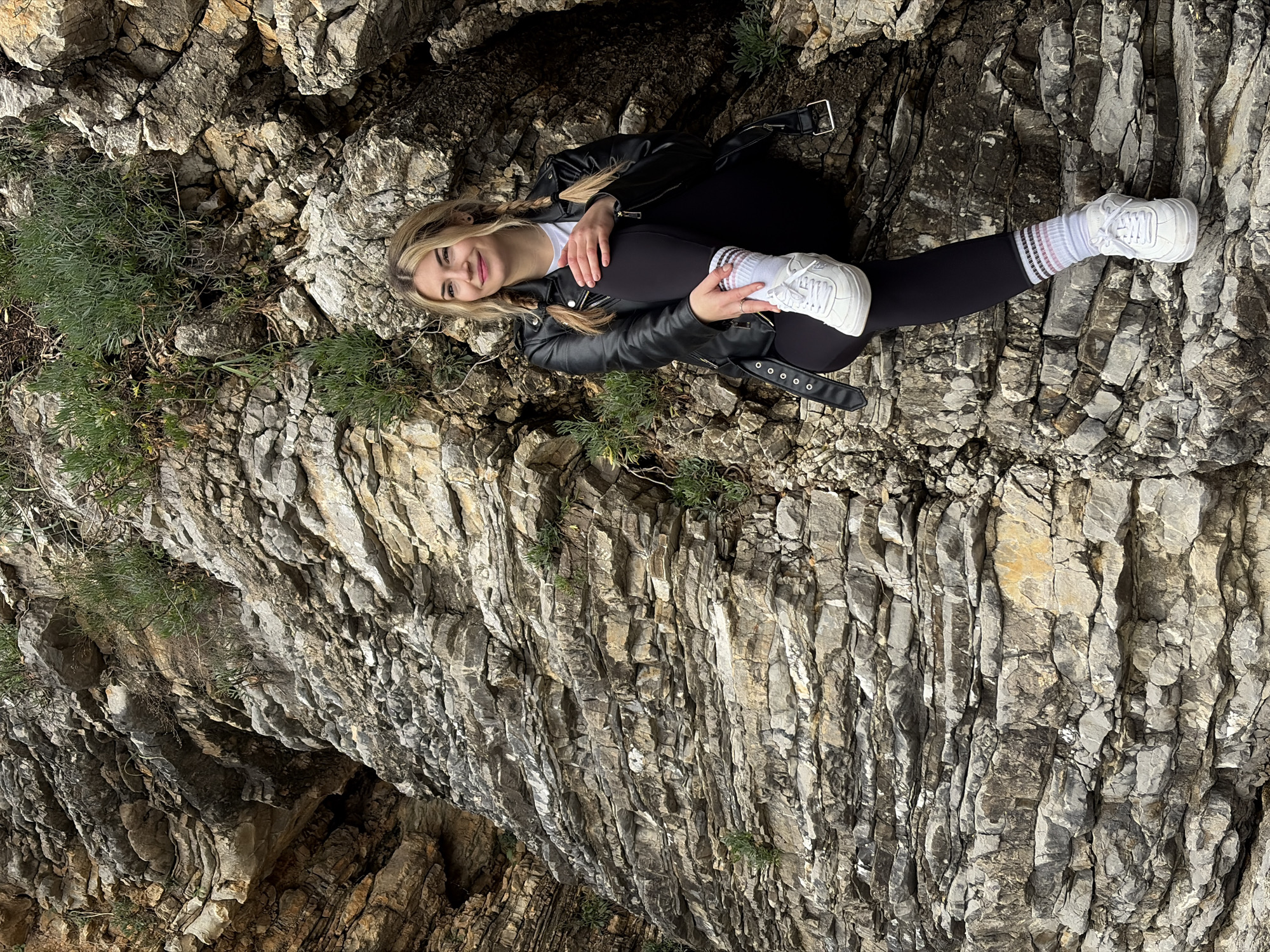 The image depicts a woman lying on a rock formation in Budva, Montenegro. She is wearing a black leather jacket, a white t-shirt, black leggings, and white sneakers with striped socks. She has blonde hair styled in pigtails. The rock formation is predominantly gray with some green vegetation growing on it. The image is taken from a slightly elevated angle.
