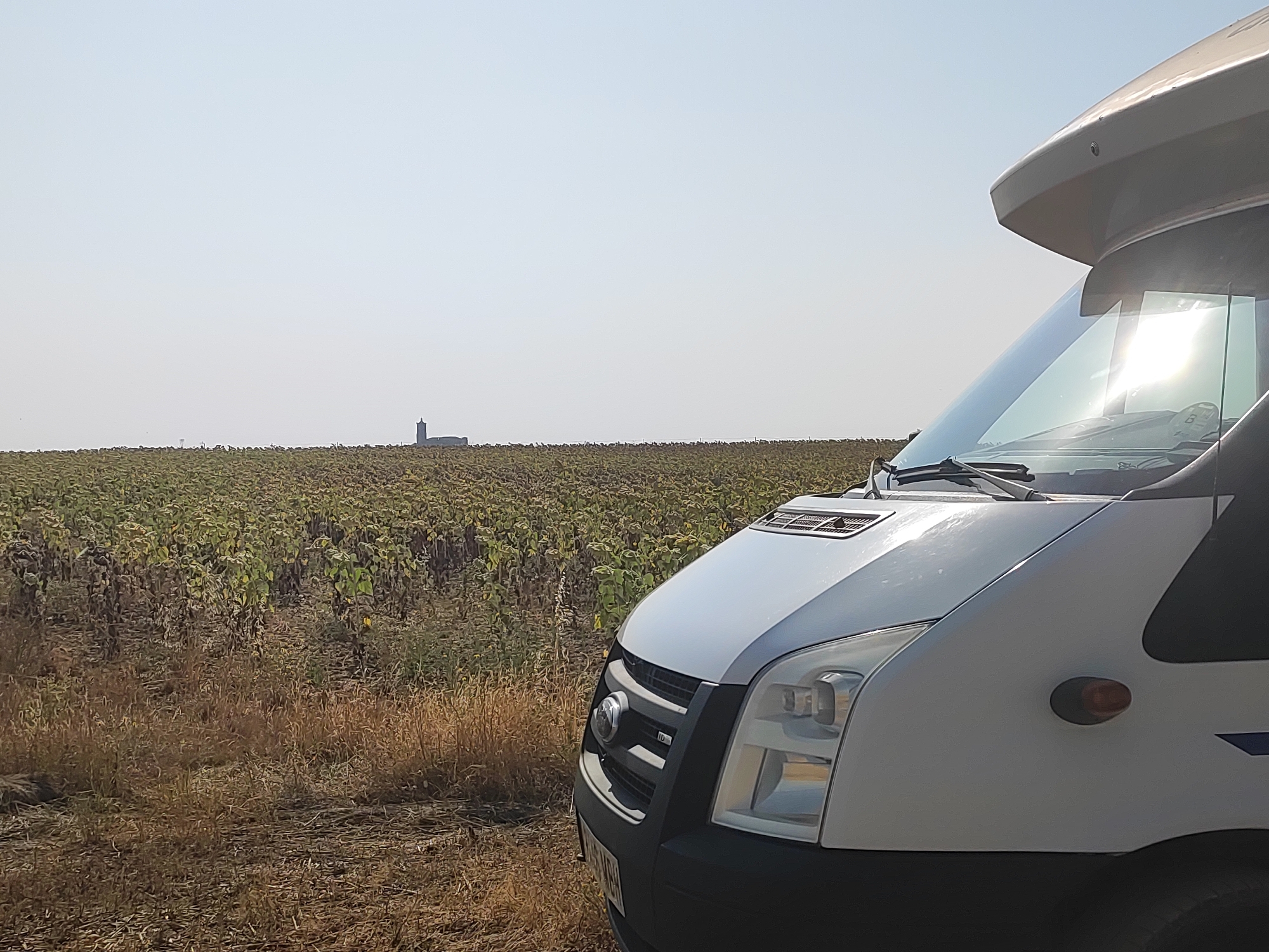 In the foreground, a white motorhome is parked on the side of a dirt road, its front right quarter visible. The vehicle's headlights, grille, and part of its windshield are in focus. The sun is reflecting brightly off the windshield. In the mid-ground and background, a large field of sunflowers stretches to the horizon. The sunflowers appear to be past their prime, with many heads drooping. In the far distance, a silhouetted structure resembling a tower or silo can be seen against the pale sky. The overall environment suggests a rural setting under a bright, possibly hazy, sky. The location is Autillo de Campos, Spain. There are no people visible in the image.