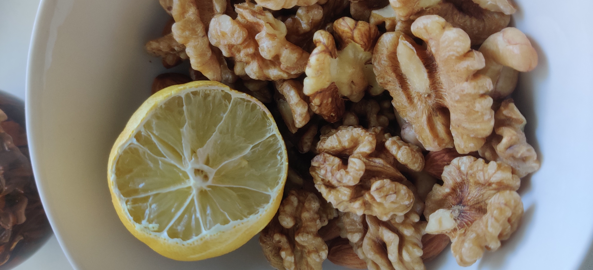 A close-up, overhead shot shows a slice of lemon resting on a pile of walnuts. The lemon slice is positioned in the upper left quadrant of the frame, its textured rind and segmented pulp clearly visible. The walnuts, in varying shades of brown and beige, fill the majority of the frame, appearing to be shelled and whole. A white bowl partially frames the subjects. The background is out of focus, showing the dark brown interior of another bowl. The scene is lit from above, casting subtle shadows. There is no indication of the city of Yekaterinburg, Russia, or any people in the image.