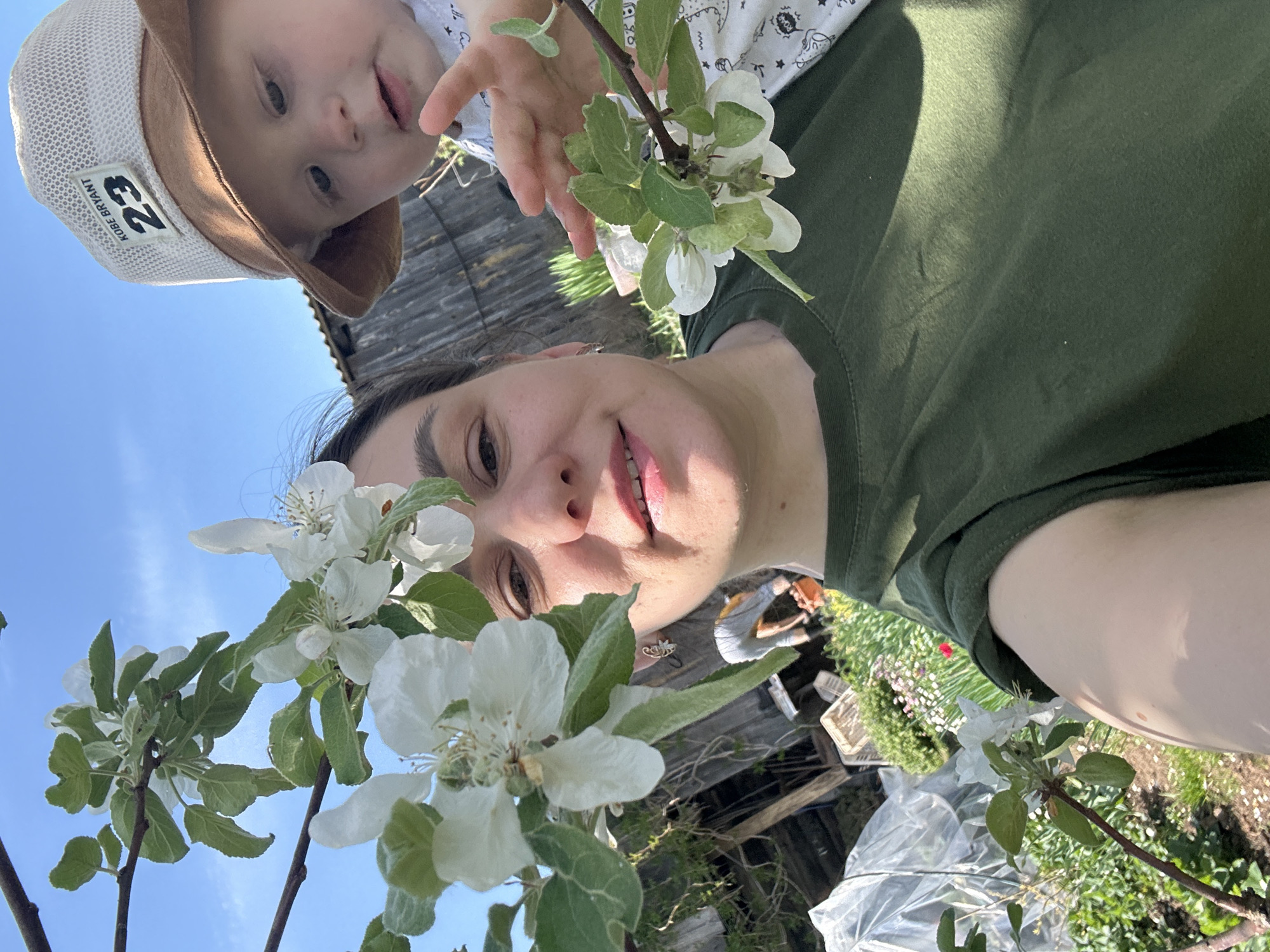 A woman and a baby are in a garden, likely in the city of Фоки, Russia. The woman is wearing a green shirt and smiling. The baby is wearing a light-colored hat with a "23" patch and a light-colored patterned shirt. Both are looking at the camera. The garden has flowering trees, possibly apple trees, with white blossoms. A wooden building is in the background. The sky is blue. There is clear plastic sheeting and other gardening items on the ground. The image appears to be taken from a low angle.