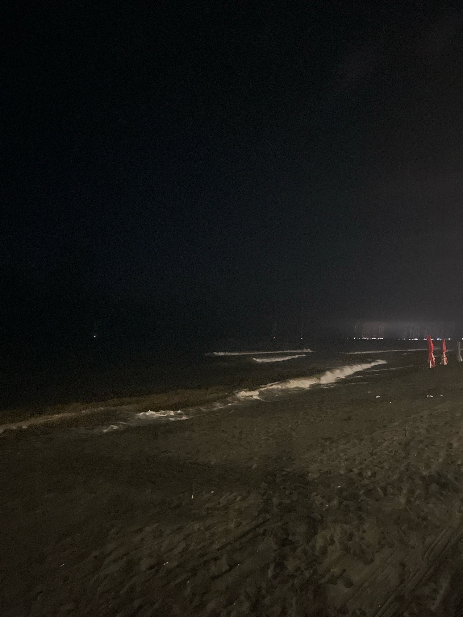 The image shows a dark, sandy beach at night in Ureki, Georgia. The sea is calm with gentle waves breaking on the shore. The sky is dark and appears overcast, with only a few faint lights visible in the distance, possibly from a town or a street. There are a few red umbrellas on the beach, suggesting that this is a popular tourist area. The overall scene is peaceful and serene, with the sound of the waves and the gentle night air creating a tranquil atmosphere.