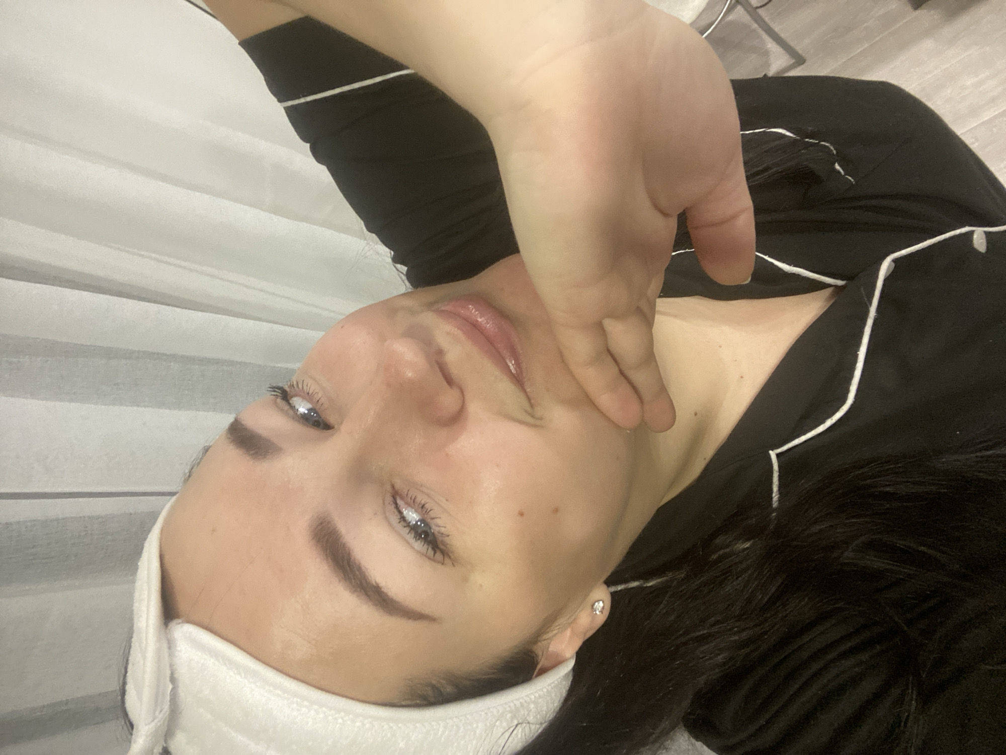 This image, taken in Sochi, Russia, features a person, likely a woman, in an indoor setting. The photograph is rotated 90 degrees clockwise, showing the person lying down with her head towards the left side of the frame, looking up. She has dark hair, light-colored eyes (blue or grey), and well-defined eyebrows.
She is wearing a white headband or hair wrap, keeping her hair away from her face, and a black top with white piping details. Her skin appears to be moisturized or has a product applied, giving it a somewhat glossy finish, especially around her lips and cheeks. Her left hand is gently placed on her jawline and cheek, suggesting she might be undergoing a facial massage or treatment, or simply posing while touching her face. A small, clear stud earring is visible in her right ear.
The background on the left consists of light-colored, possibly white, drapes or blinds with horizontal folds. On the right side, parts of a dark surface (possibly clothing or hair) and a metallic furniture leg are visible against a light-colored floor. The overall impression is one of relaxation or a self-care activity. There is no visible text in the image.
