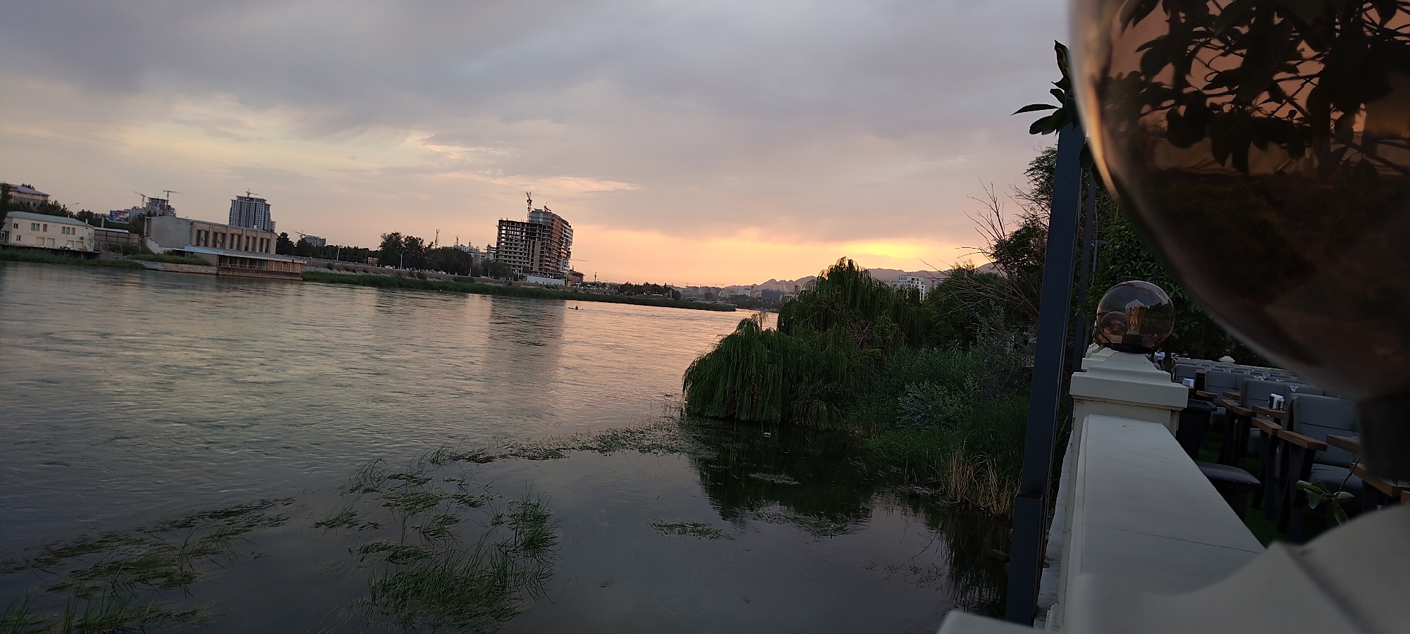 The image depicts a wide, calm river flowing through an urban landscape during sunset. In the foreground, to the right, a white railing is visible, with a decorative spherical light fixture on a pillar and a large, reflective sphere obscuring part of the view. Beyond the railing, rows of empty outdoor seating are arranged. The river's surface reflects the muted tones of the sky and the surrounding environment. On the opposite bank, buildings are visible, including a multi-story structure under construction with cranes. Further in the distance, hills form the horizon under a sky transitioning from dusky clouds to a warm, orange glow from the setting sun. The scene suggests a waterfront establishment or public space in Khujand, Tajikistan, possibly a restaurant or cafe overlooking the Syr Darya River.