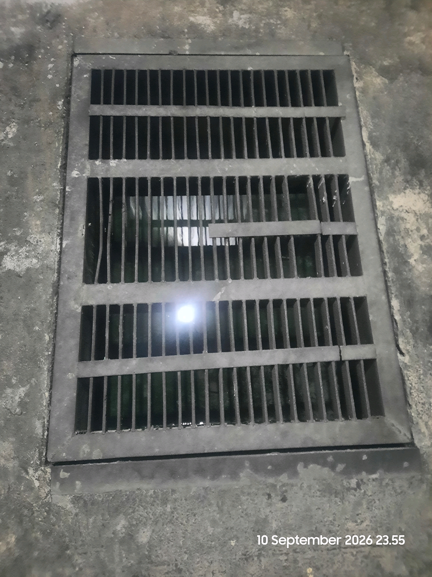 The image shows a metal grate covering a drain, likely on a concrete surface. The grate is rectangular with evenly spaced vertical bars and horizontal supports. There appears to be water or a dark substance beneath the grate, and a bright light source is visible reflected in the water. The surrounding area is a rough, textured concrete, possibly a sidewalk or roadway. In the bottom right corner, a timestamp reads "10 September 2026 23.55". The image does not contain any information that would indicate its location in Gending, Indonesia.