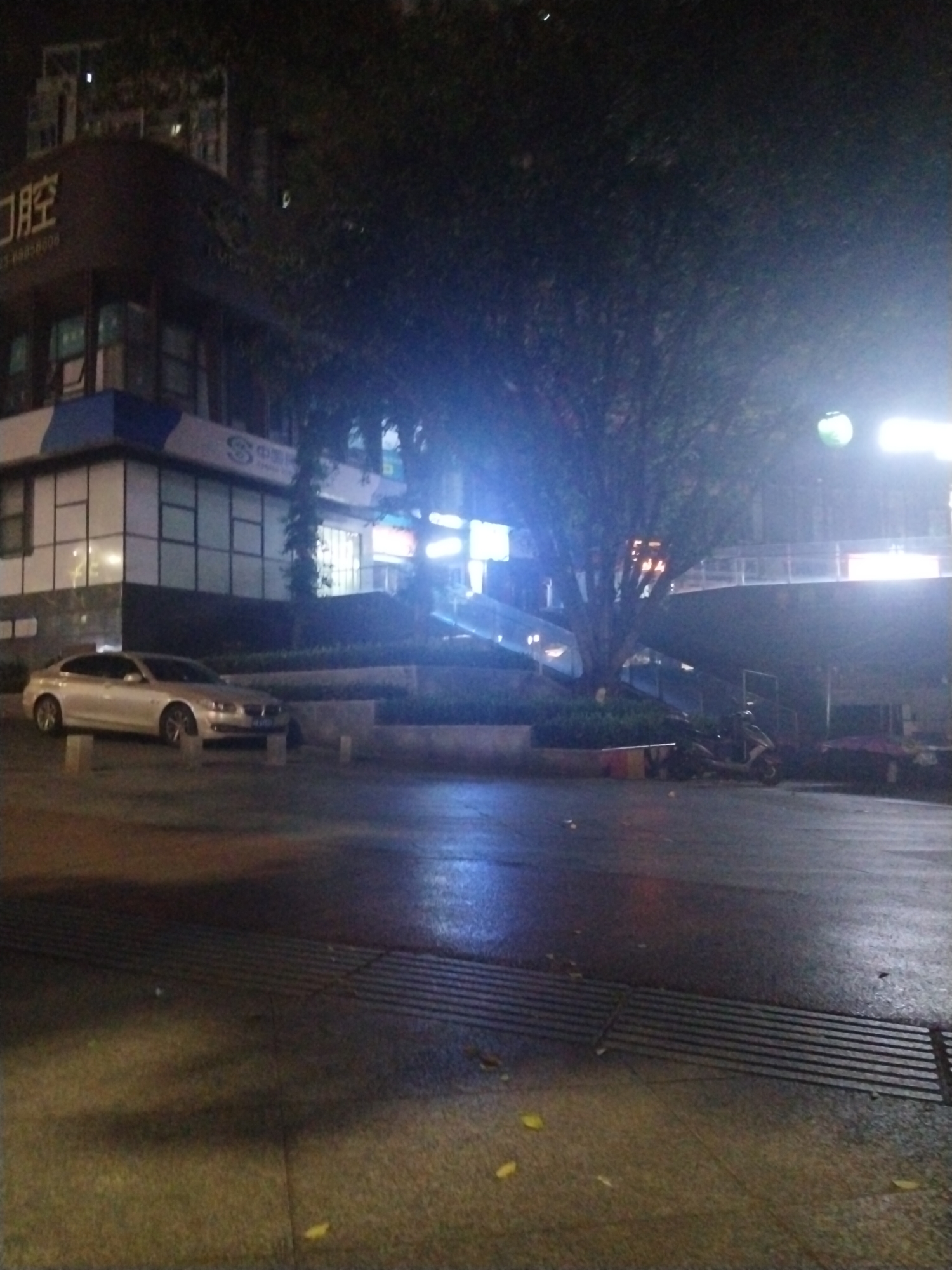 The image depicts a street scene at night in Jiulongpo District, China. A light-colored car is parked on the left side of the frame near a multi-story building with illuminated windows. In the center, a large tree obscures the view. A pedestrian walkway with a ramp and stairs leads up to a higher level. To the right of the walkway, a scooter and a vendor's setup are visible. The street is wet and reflects the surrounding light. A metal grate is visible in the foreground of the scene. The overall ambiance is dark, with light sources scattered throughout the scene.