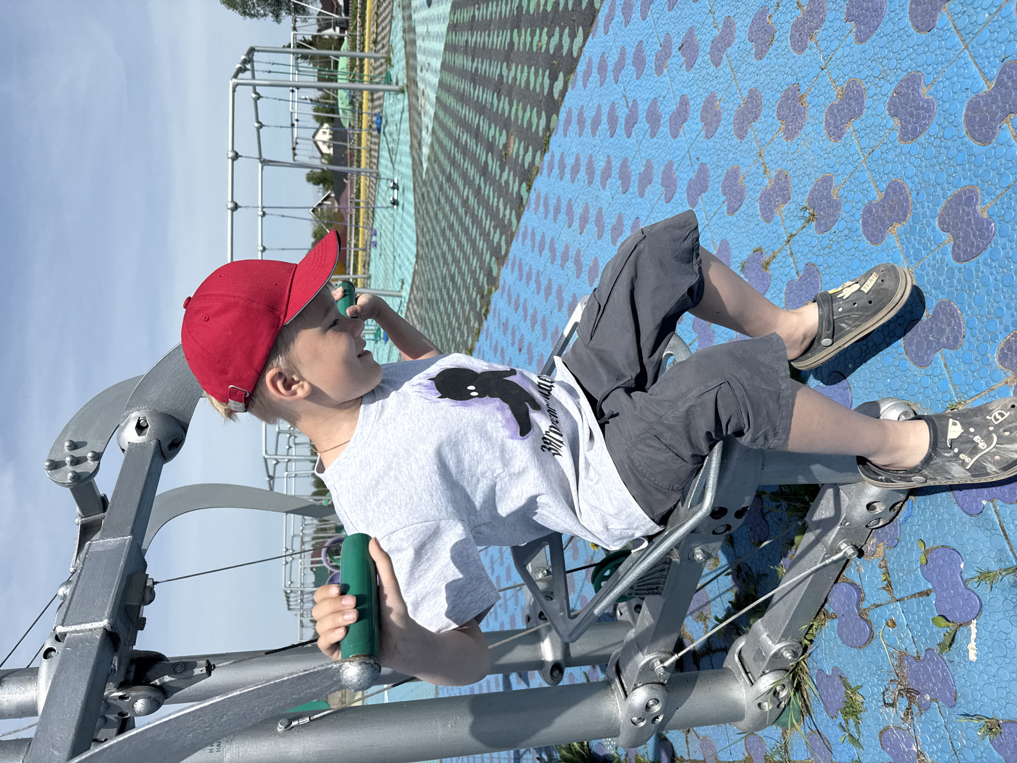 A young boy with short blond hair wearing a red baseball cap, a gray t-shirt with a black cartoon print, and gray shorts sits on a gray metal contraption on a playground in Синьково, Russia. He is holding a green handle with a silver ball at the end. The playground has a blue surface with purple patterns and a green and black striped area. In the background, there is a gray metal structure, some houses, and trees.