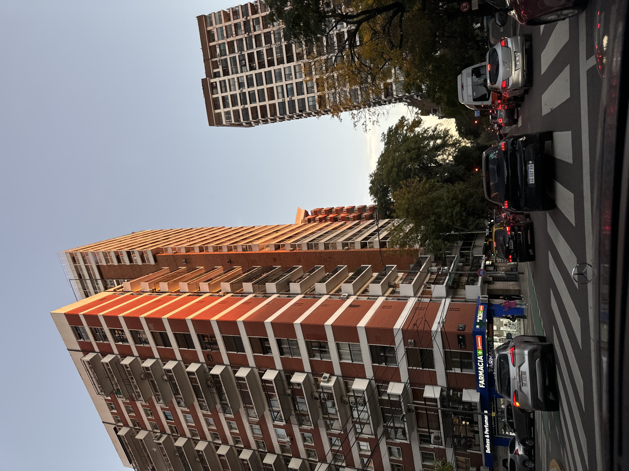 The image captures a street scene in Buenos Aires, Argentina, featuring two high-rise buildings. The foreground building is characterized by a red brick facade with white horizontal stripes and numerous balconies. A pharmacy ("FARMACIA + GUINESTAR") is located at the building's base, with visible signage. Cars are stopped at a zebra crossing in the foreground, with visible license plates. The second building is visible at the top and is a bit farther in the distance and has many windows. The sky is clear.