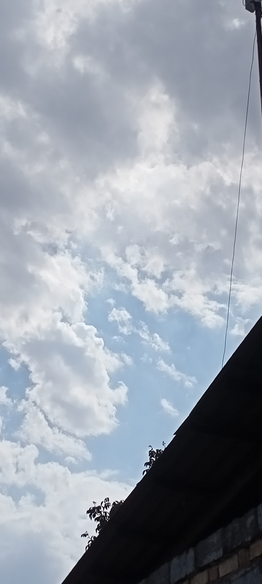 The image captures a view of a cloudy sky with a dark roofline in the foreground. A dark pole with a fixture at its top extends vertically on the right side of the image. The roof appears to be made of corrugated material, and below it, a brick wall is visible. Some foliage, likely from a tree, is silhouetted against the sky at the edge of the roofline. The scene is set outdoors in the city of Sokuluk, Kyrgyzstan.