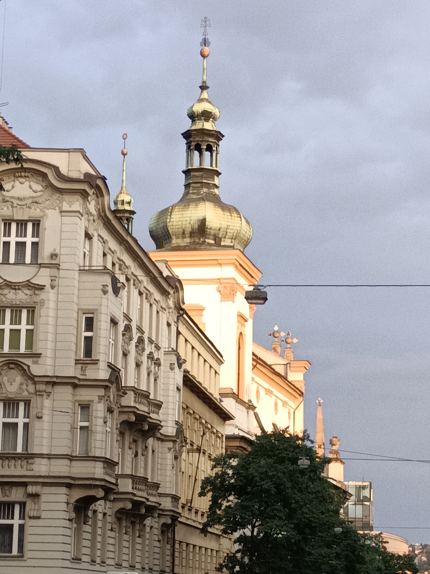 This is a medium shot of the facade of buildings in Prague, Czechia, against a cloudy sky. The focus is on the architectural details of the buildings, which are in a Baroque style. The buildings are tall and narrow, with ornate decorations around the windows and balconies. The prominent building on the right has a large, green dome with a spire topped by a cross. There is a dark green tree in the foreground, obscuring some of the lower parts of the buildings. Power lines are visible across the frame. The lighting suggests it is late afternoon or early evening.