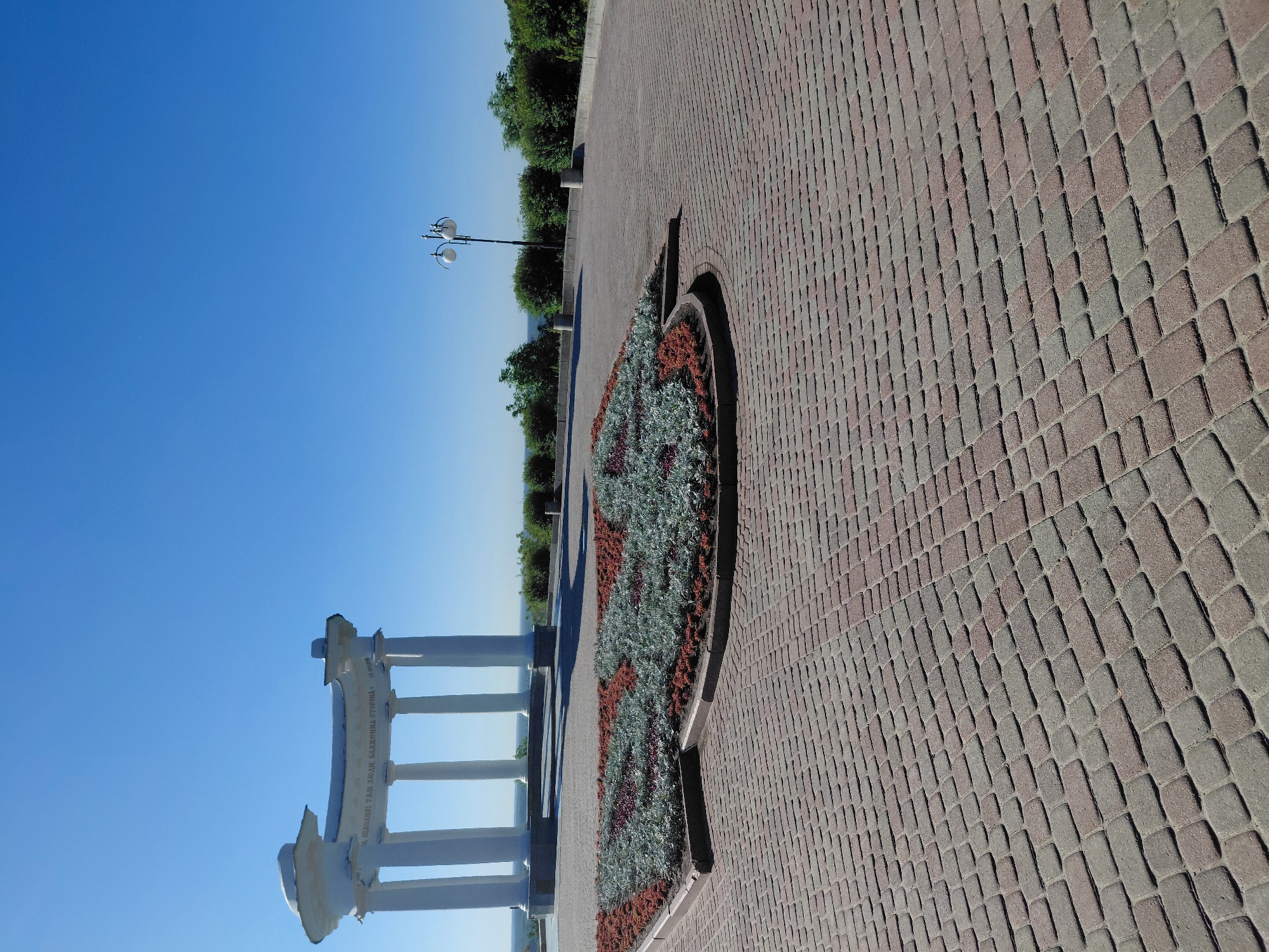 The image, taken in Poltava, Ukraine, depicts a serene outdoor scene under a clear blue sky. The foreground features a neatly paved walkway constructed from reddish-brown and grey interlocking bricks, arranged in a distinctive diagonal pattern. Adjacent to this paving is a crescent-shaped flowerbed, meticulously planted with a variety of foliage in shades of green, grey-green, and reddish-brown, which adds vibrant texture to the scene.
Further into the mid-ground, a prominent white balustrade with multiple vertical columns creates a decorative barrier, suggesting a viewpoint or an edge of a raised area. Visible on the curved, upper section of this structure is Cyrillic text, which reads: "І.П. ОВСЯННІКОВЪ ТАМ АРХІТ. БАЖЕНОВЪ ВТОРОЙ". This inscription identifies the structure as the White Arbor (Bila Besidka) in Poltava, a notable landmark.
A classic black streetlamp with two lights stands among a line of lush green trees that form a natural boundary in the mid-ground. The background is dominated by a vast expanse of clear, vibrant blue sky, indicating bright, sunny weather. No people are visible, contributing to a tranquil and contemplative atmosphere. The strong, even lighting suggests it is daytime, likely morning or early afternoon. The scene conveys a sense of peaceful urban landscaping combined with historical significance.
