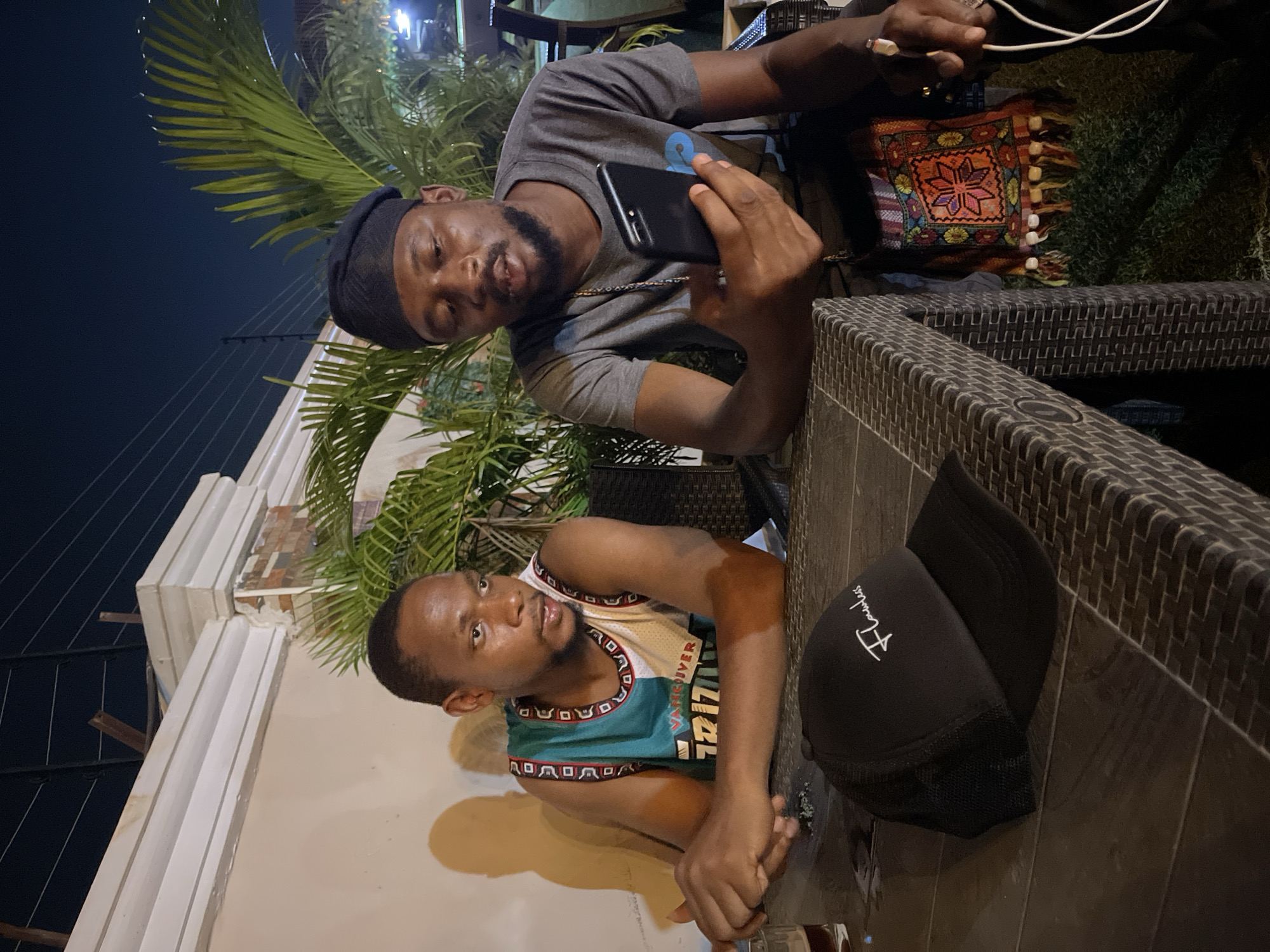 The image captures two men in what appears to be an outdoor or semi-outdoor dining or lounge area at night, likely in Abuja, Nigeria.
The man on the left is seated, wearing a grey t-shirt, a dark beanie, and a gold chain. He is holding a black mobile phone in his right hand, seemingly looking at the screen or taking a photo/video. His left arm rests on a dark, possibly rattan, table. Behind him is a vibrant, multi-colored decorative cushion with intricate patterns and tassels, resting against another dark chair. A white charging cable is visible near his right hand.
The man on the right is also seated at the same table, looking towards the camera or slightly to the left. He is wearing a teal and white sleeveless top with "Vancouver" text visible on the front. His hands are clasped on the table in front of him. A black cap with white embroidered text reading "Flawless" is placed on the table to his right.
The setting is illuminated by artificial light sources, casting shadows and highlighting the subjects. In the background, lush green palm plants are visible, suggesting a warm climate. The furniture consists of dark, possibly rattan-style chairs and tables. The overall atmosphere appears casual and relaxed, indicative of a social gathering during the evening.