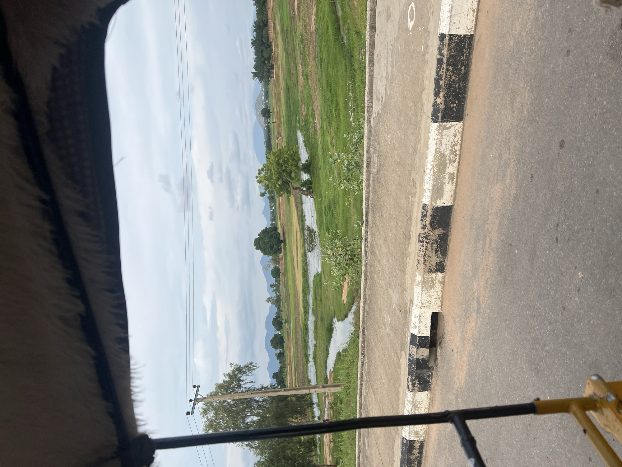 The image, captured from the perspective of a moving vehicle, likely a rickshaw due to the visible yellow and black metal frame and the furry interior lining of the roof, shows a rural or suburban landscape. The foreground features an asphalt road with a white and black striped curb, indicating a maintained pathway. Beyond the curb, a body of water, possibly a river or a flooded area, meanders through a lush green field dotted with trees. In the distance, a range of mountains can be seen under a cloudy sky. A utility pole with power lines stretches across the frame, suggesting the presence of electricity infrastructure. The overall scene depicts a serene, natural environment with signs of human habitation and infrastructure. This scene is consistent with the outskirts of Jalingo, Nigeria, where such landscapes are common.