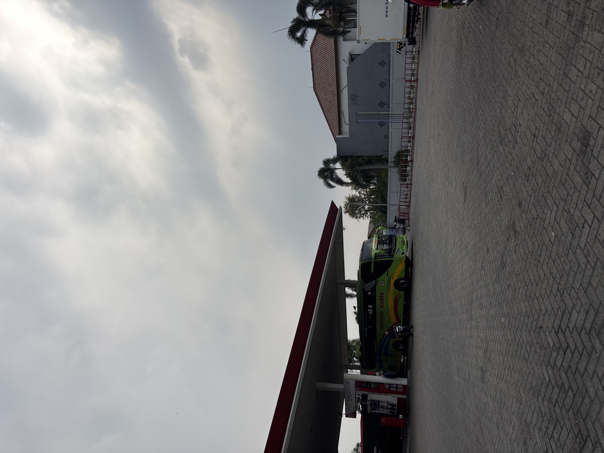 This is a wide shot of a gas station in Bekasi, Indonesia, under a cloudy sky. A large, green and yellow bus with the text "GUNUNG HARTA" is parked under the red-trimmed canopy of the gas station. A person is seated on a motorcycle in front of the bus, facing left. To the left of the bus, a red gas pump is visible. Behind the gas station, there is a white building with a red-tiled roof, surrounded by a grey wall and some palm trees. A white truck with "www.be" visible on its side is parked to the right. The ground is paved with dark grey interlocking bricks.