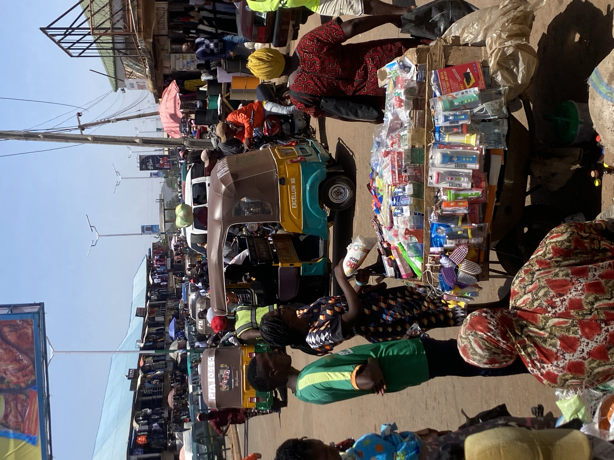 The image captures a bustling street scene in Bukuru, Nigeria. A line of auto-rickshaws, locally known as keke, are parked along the road, including one marked "EXCELLON X6." People are moving about, with some appearing to be vendors displaying their wares. One vendor's table showcases an array of packaged items, possibly household goods or small electronics. In the foreground, a young woman in a patterned dress holds a package labeled "MOP," interacting with a young man in a green shirt. Overhead, power lines crisscross the sky. Billboards and commercial signs can be seen in the background, indicating a lively commercial area. The presence of makeshift stalls and vendors suggests a marketplace atmosphere.