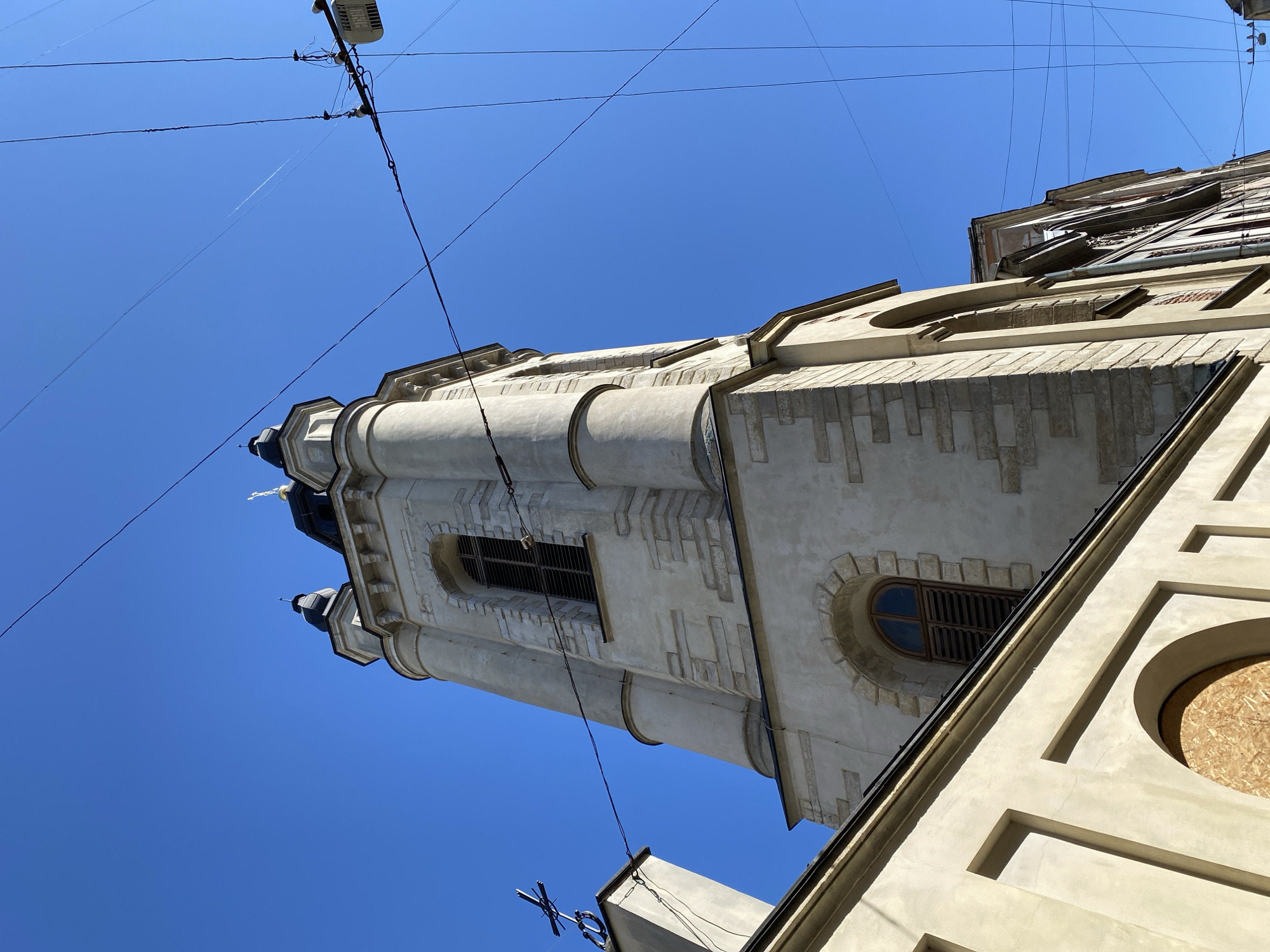 The image shows a view from below of a stone church tower with multiple levels and spires, against a clear blue sky. The tower is light-colored, with architectural details such as arched windows with shutters, decorative stone accents, and cornices. At the top of one spire is a cross. A part of the church's lower structure, with arched windows, is visible in the foreground on the left. Overhead, a complex network of power lines and a streetlight are present, crossing the sky and partially obscuring the upper portion of the tower. One of the windows on the lower structure has been boarded up with plywood. The overall impression is of an old, historic building in an urban environment, possibly in a well-established part of Lviv, Ukraine, given the architectural style and the presence of overhead wires common in older city centers.