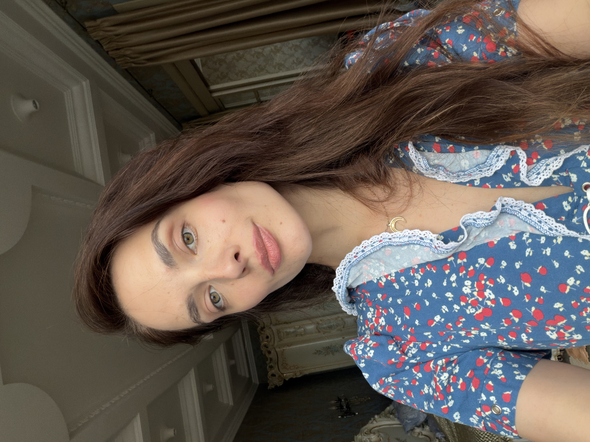 This media file is a portrait of a young woman with long, dark brown hair. She is wearing a blue dress with a red and white floral pattern and white lace trim around the neckline. A gold necklace with a crescent moon pendant is visible around her neck. The woman is looking directly at the camera with a neutral expression. Her eyes are green, and she has light skin with some freckles.
The background appears to be an indoor setting, possibly a bedroom, with decorative molding on the walls and a chandelier visible in the background. The lighting suggests it could be daytime, possibly late afternoon, as there is natural light coming from a window. The overall mood is intimate and personal, given the close-up nature of the shot and the direct gaze. There is no visible text in the image. The provided location context is Ufa, Russia.