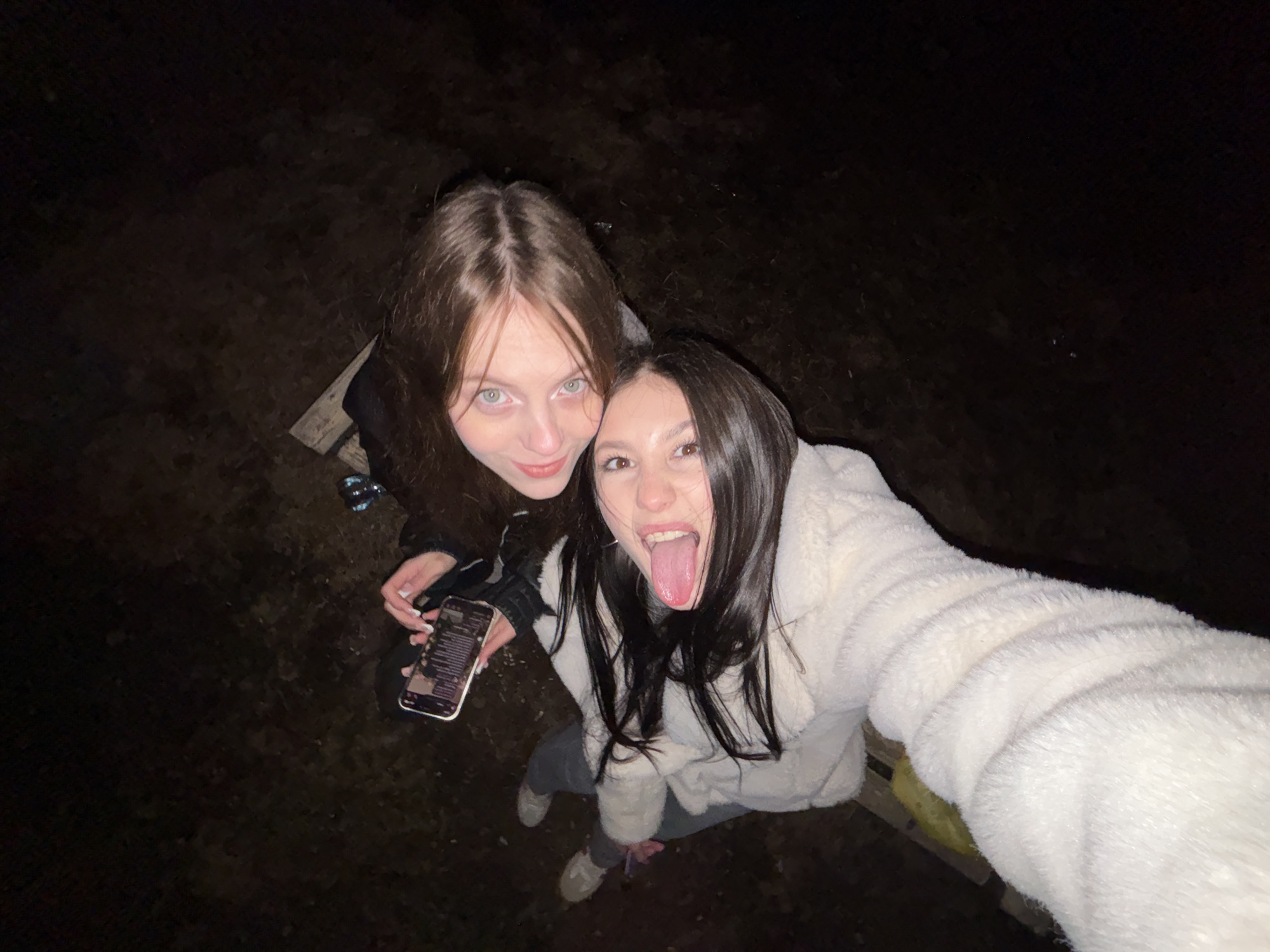 Two young women are taking a selfie outdoors at night. The woman on the left has light brown hair and green eyes, and is holding a phone with a lit screen. The woman on the right has long dark hair and is sticking her tongue out. Both women are wearing jackets. The scene is dimly lit, and the ground appears to be dark and uneven. The specific location in the city of Фоки, Russia cannot be determined from the image.