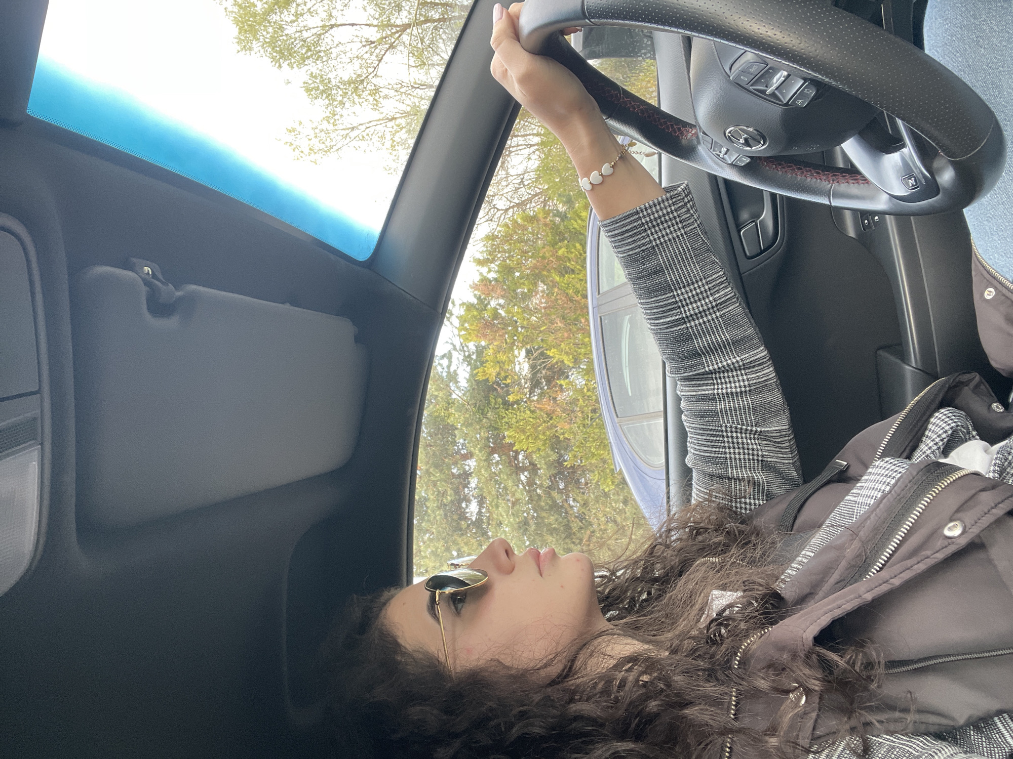 The media file captures a relaxed moment inside a car, featuring a young woman who is likely reclining in the driver's or front passenger seat, with the image rotated 90 degrees counter-clockwise. The setting is Sofia, Bulgaria.
The main subject is a woman with long, dark, curly hair, wearing aviator-style sunglasses. She is looking upwards, presumably through the car's windshield at the sky and surrounding trees. Her attire includes a dark puffer vest worn over a grey and white plaid top or jacket. On her right wrist, she wears a delicate bracelet with several small, white, heart-shaped beads. Her right hand is casually placed on the car's black leather steering wheel, which features red stitching, controls, and a visible Mazda logo in the center. A button with the letter "N" is also discernible on the steering wheel.
The car's interior shows elements like the sun visor and part of the dashboard. Outside the vehicle, bright daylight illuminates green trees and what appears to be a clear or lightly overcast sky, indicating it is daytime, likely late morning or early afternoon. The overall impression is one of calm and relaxation, perhaps during a stop or a break from driving.