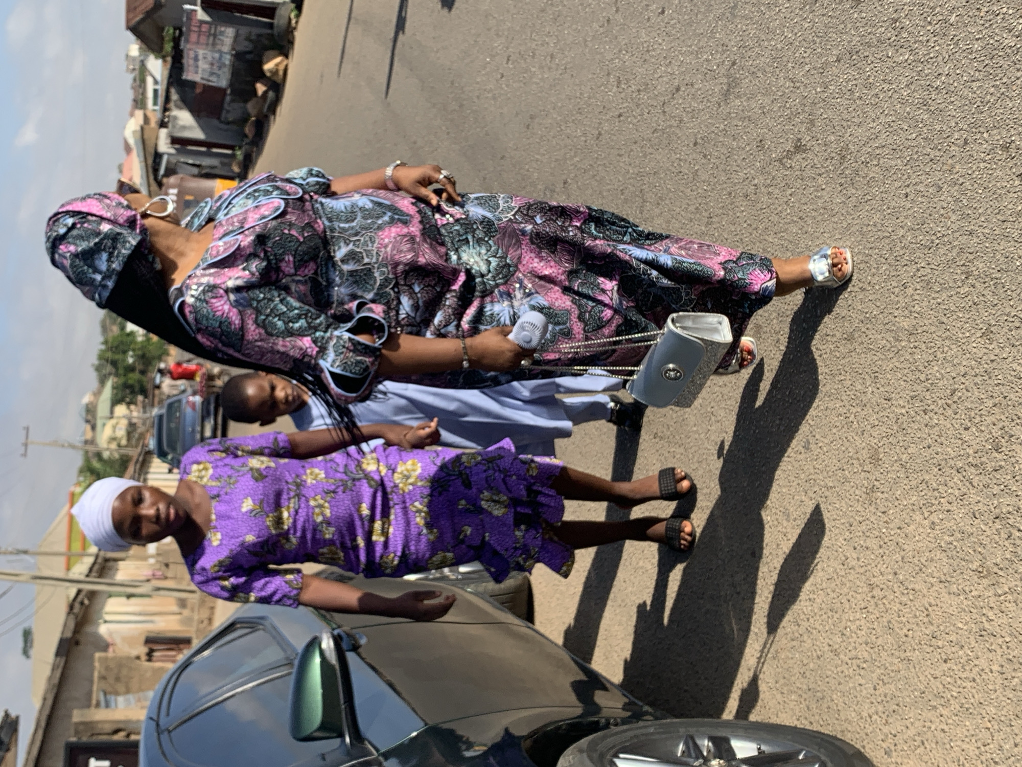 The image captures a street scene in Jos, Nigeria. A woman in a long, colorful, patterned dress and matching headwrap stands on the roadside. She is holding a small, white fan and carrying a light-colored purse with a chain strap. Beside her, a younger girl wearing a purple, floral dress and a white head covering stands near a parked car, a dark-colored sedan. Another person, wearing a light blue dress, is partially visible between them. The background features buildings and structures typical of an urban environment, with sunlight casting shadows on the street.