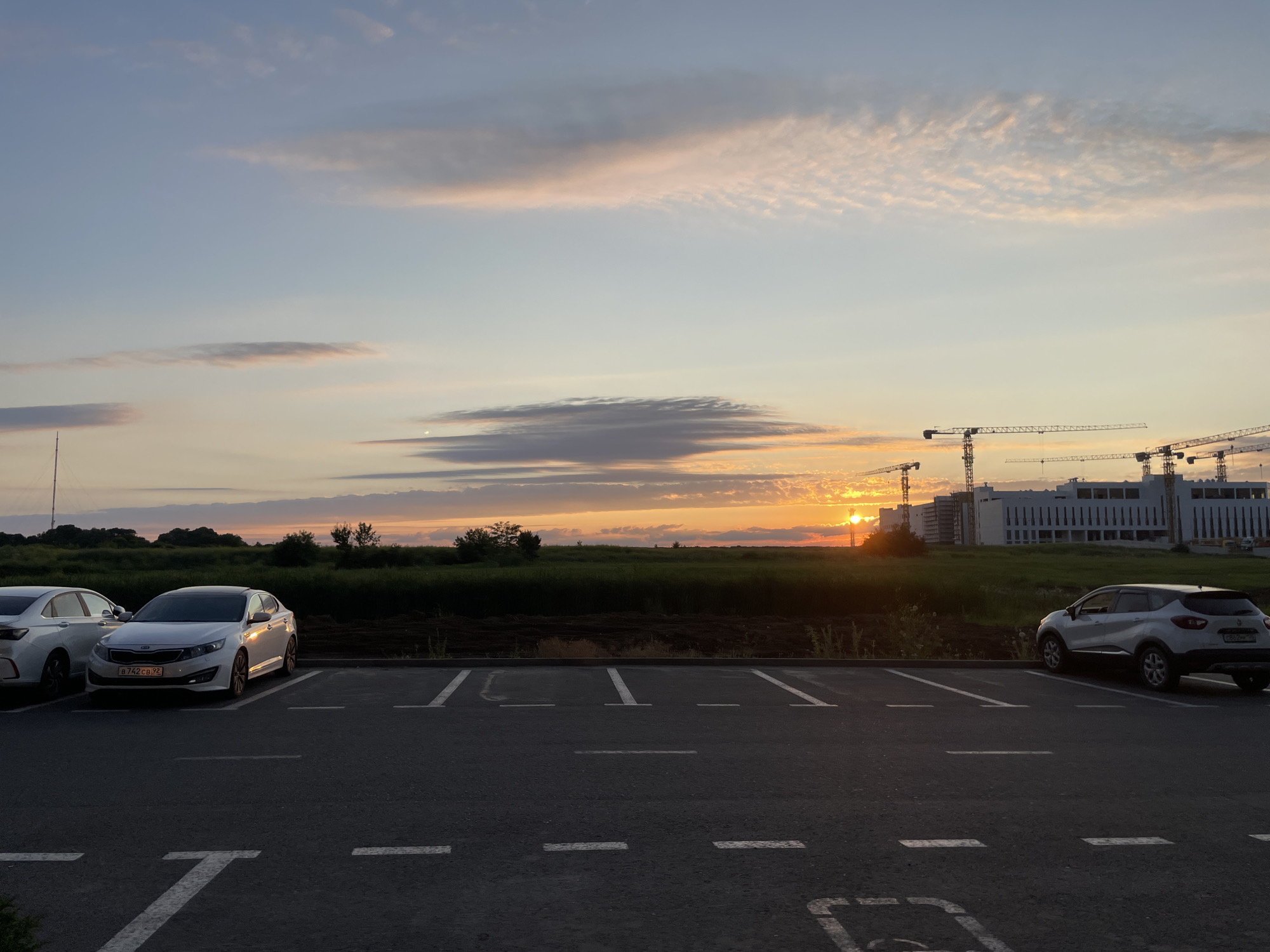 The image depicts a parking lot at dusk, likely in Stavropol, Russia, with a warm sunset casting a golden hue across the sky. Several construction cranes are visible in the distance, alongside modern buildings that appear to be under construction. The foreground shows a dark asphalt parking lot with white painted lines marking the parking spaces. Two cars are parked: a white sedan on the left and a white crossover SUV on the right. A grassy field separates the parking lot from the construction site, with trees and vegetation lining the horizon. The sky is adorned with streaks of pink and orange clouds, indicating the sun is setting behind the buildings.